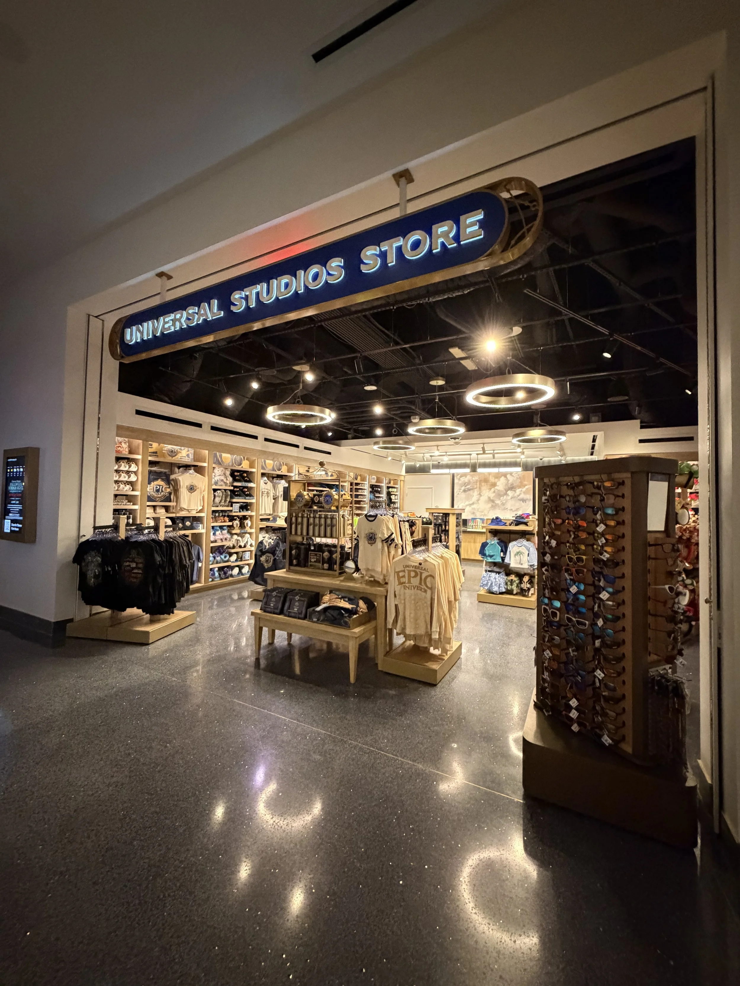 Entrance to Universal Studios Store with shelves of merchandise, including apparel, hats, sunglasses, and souvenirs, under bright round and linear ceiling lights.