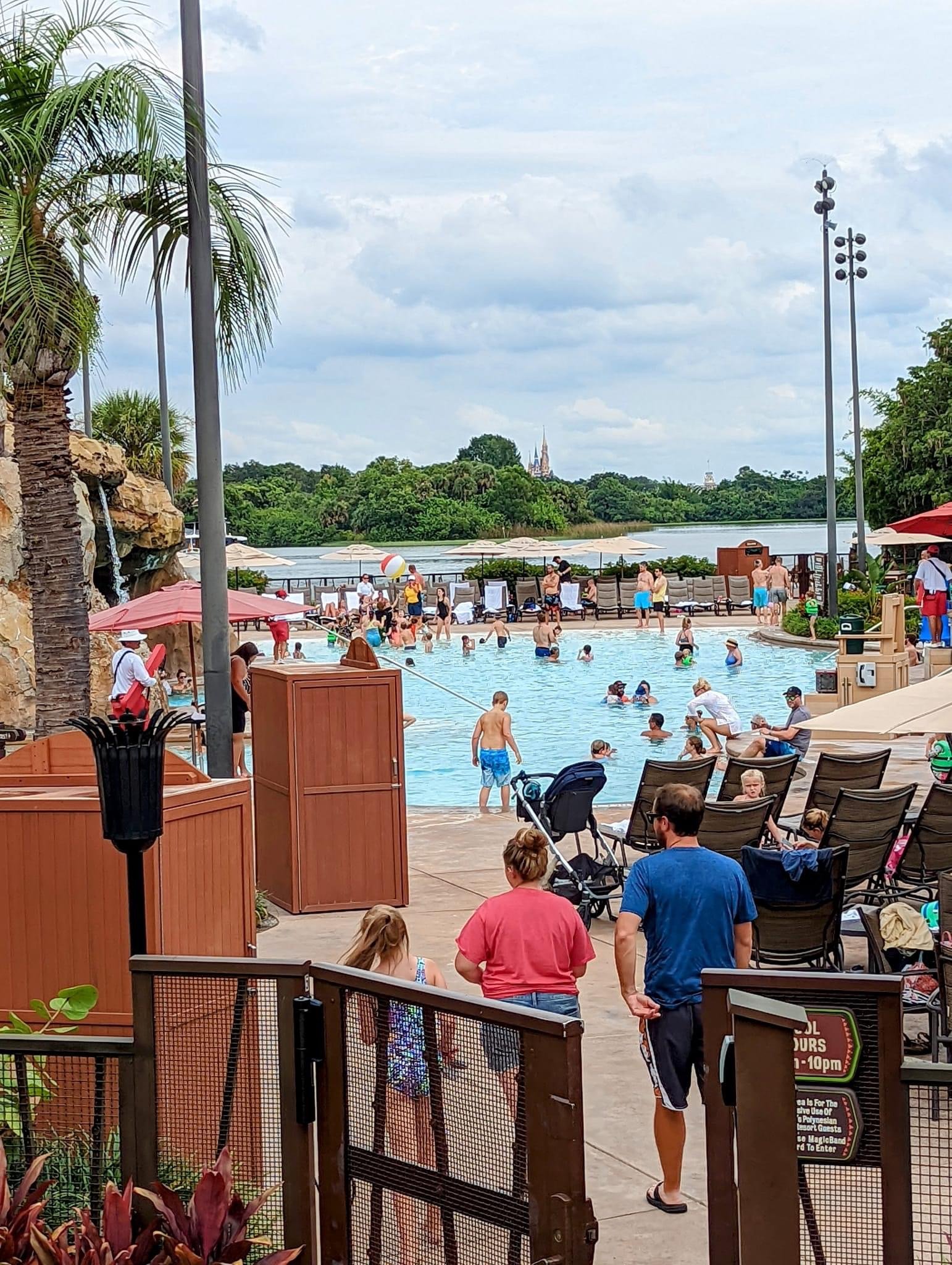 Crowded outdoor swimming pool with people swimming, relaxing, and playing, surrounded by lounge chairs, umbrellas, and greenery, with a river and trees in the background.