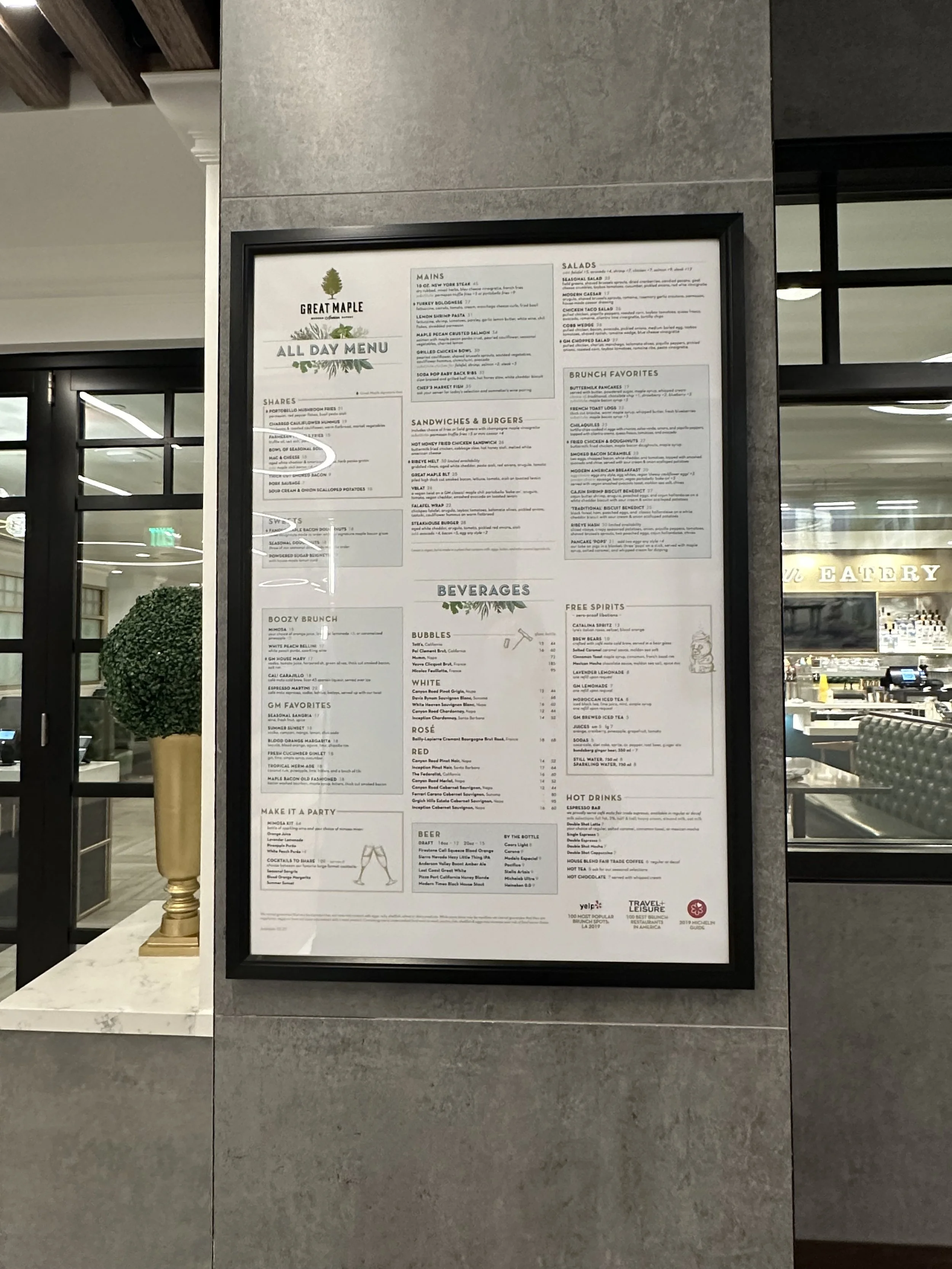 Framed menu at a restaurant called Great Maple, displaying various sections including mains, salads, sandwiches & burgers, brunch favorites, beverages, and more, with a plant in a gold vase nearby.