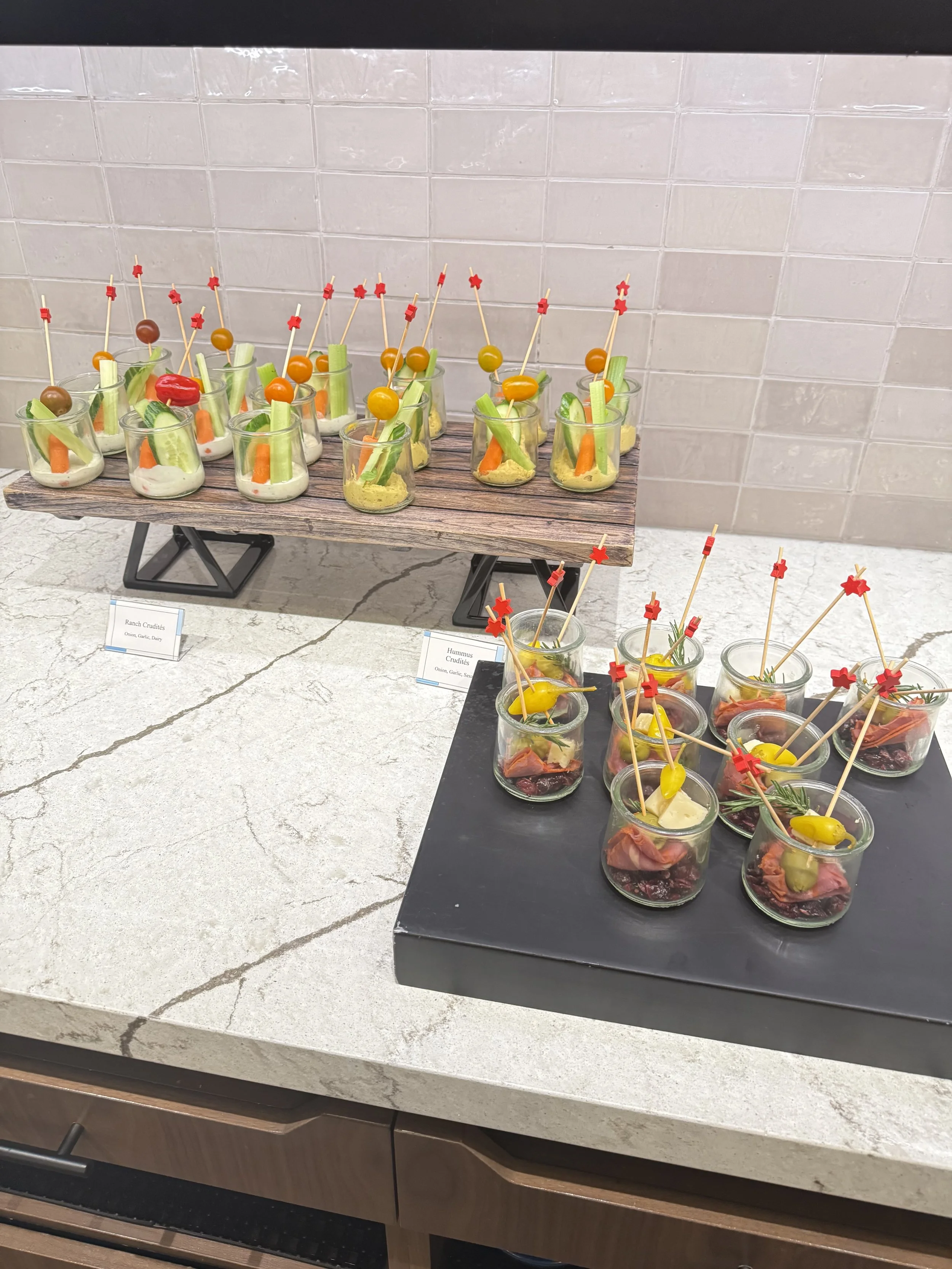 Assorted appetizers in small glasses arranged on black and wooden display stands, featuring vegetables, cheese, and cured meats with skewers and decorative picks.
