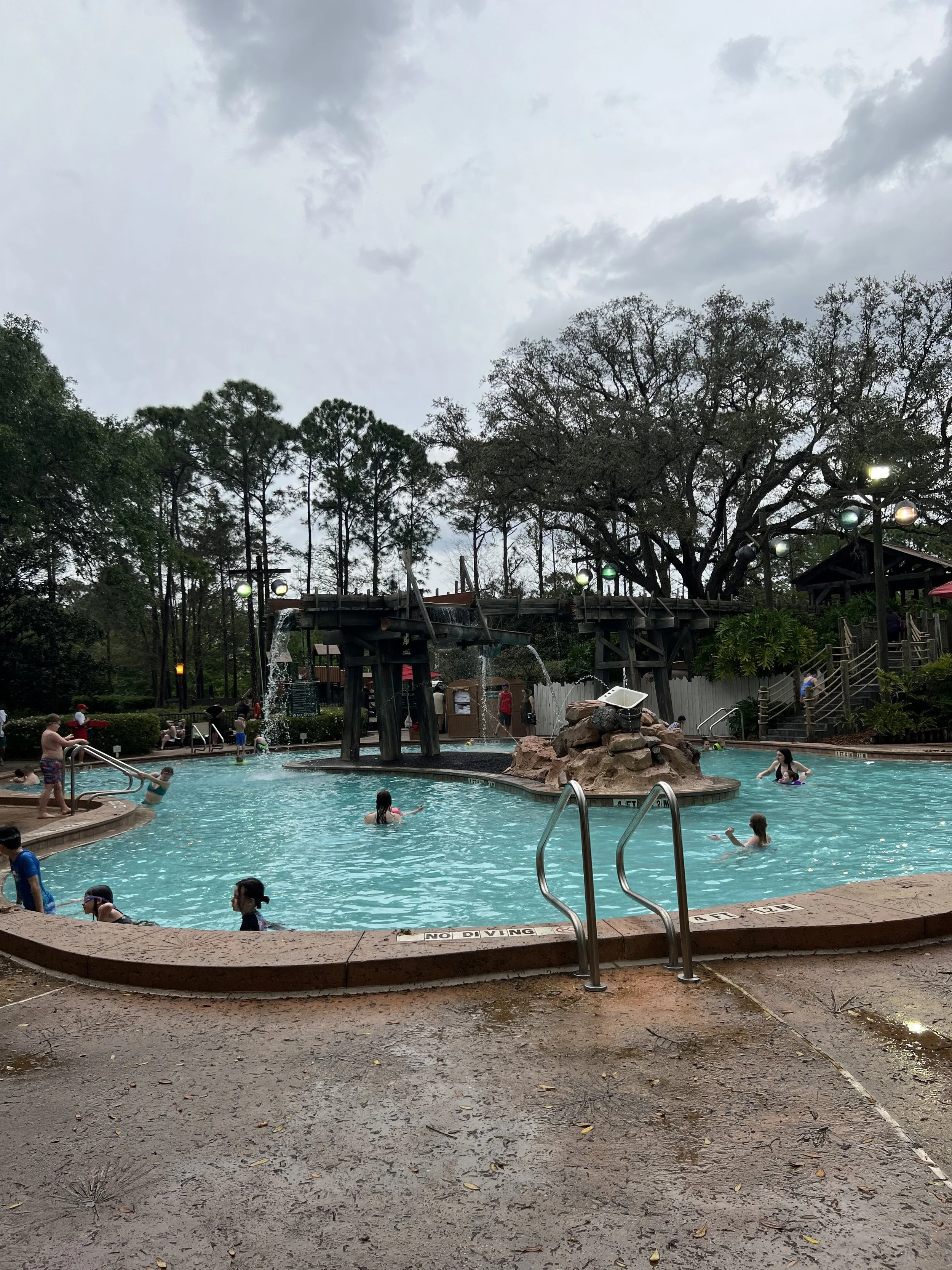 A crowded outdoor swimming pool area with people swimming and relaxing, surrounded by trees under a cloudy sky.