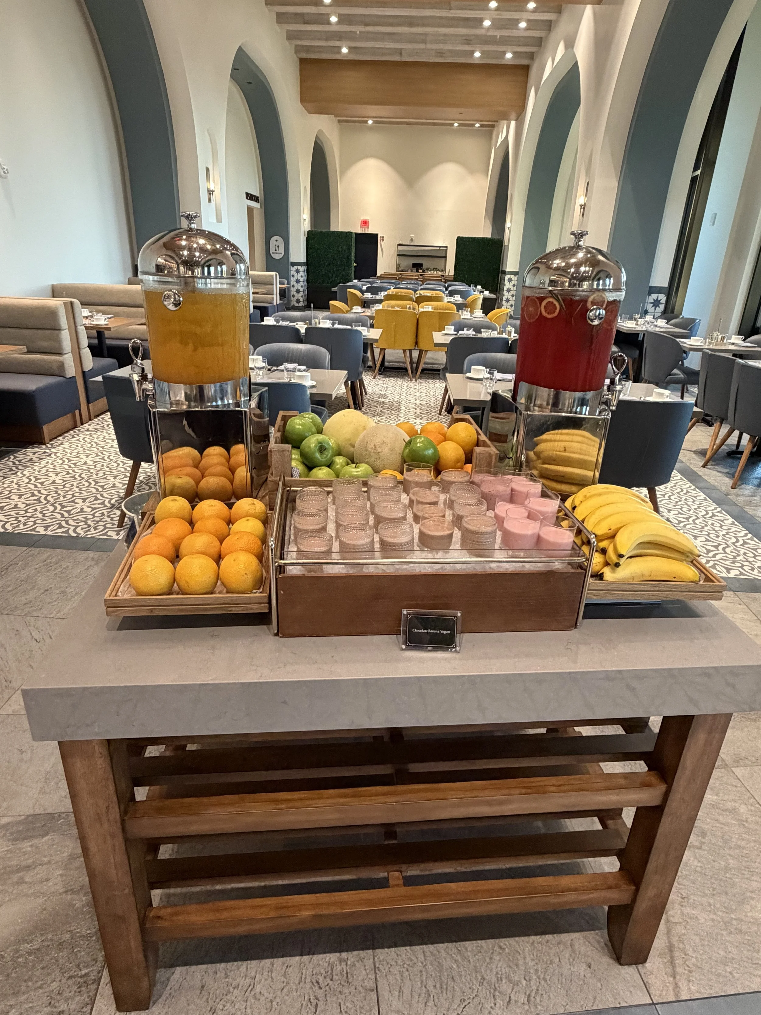 A juice and fruit station in a restaurant, with two large dispensers of orange and red drinks, surrounded by fresh fruits like oranges, lemons, apples, melons, and bananas, along with small glass containers of pink yogurt or pudding, on a wooden tabl