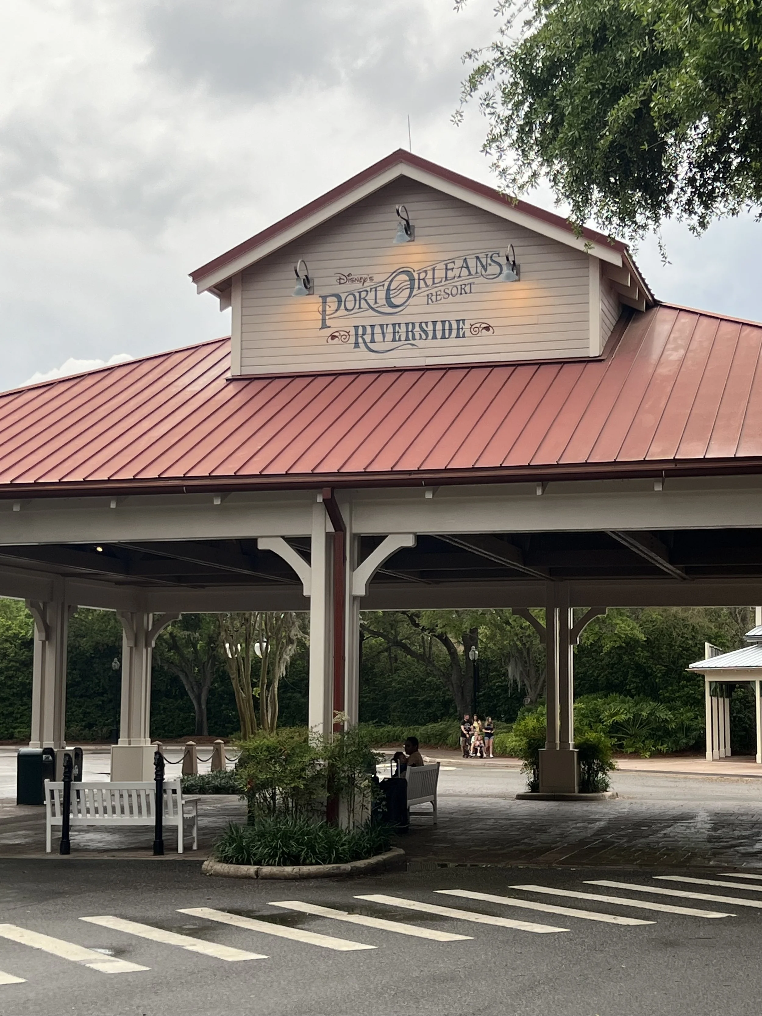 Entrance sign for Disney's Port Orleans Resort Riverside with a red metal roof and white trim, surrounded by greenery and a partly cloudy sky.