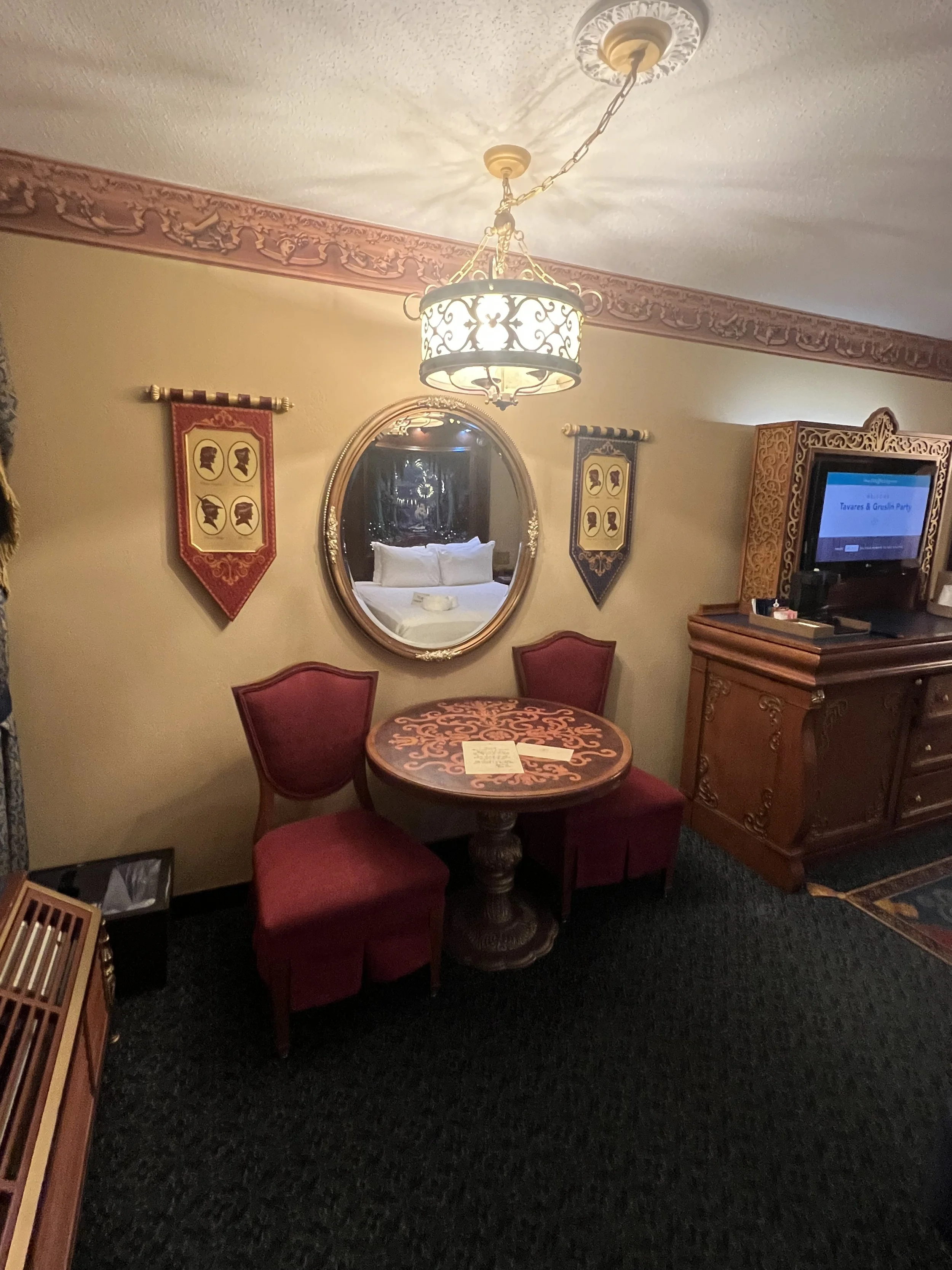 A cozy hotel room with a small round wooden table with decorative patterns, flanked by two upholstered chairs with red fabric. An ornate wall mirror is centered on the wall above the table, flanked by two decorative banners with profile silhouettes. 