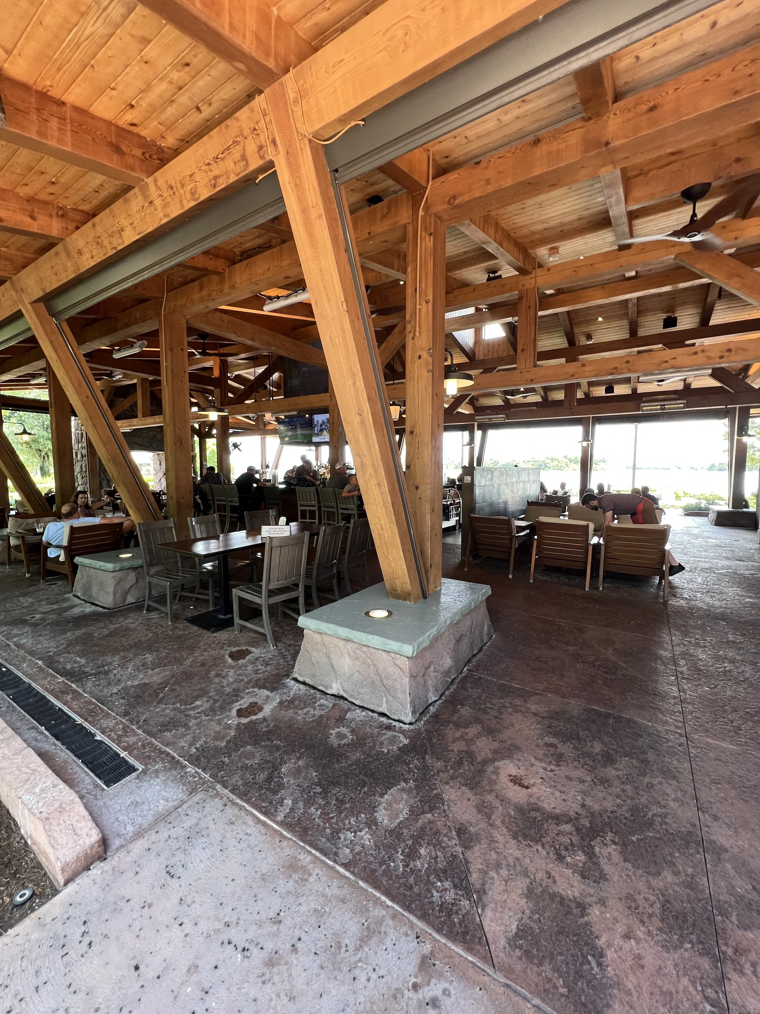 Interior of a rustic restaurant or cafe with wooden beams and ceiling, stone and concrete flooring, and several tables and chairs, some occupied by people. Large windows outline the background, allowing natural light to fill the space.