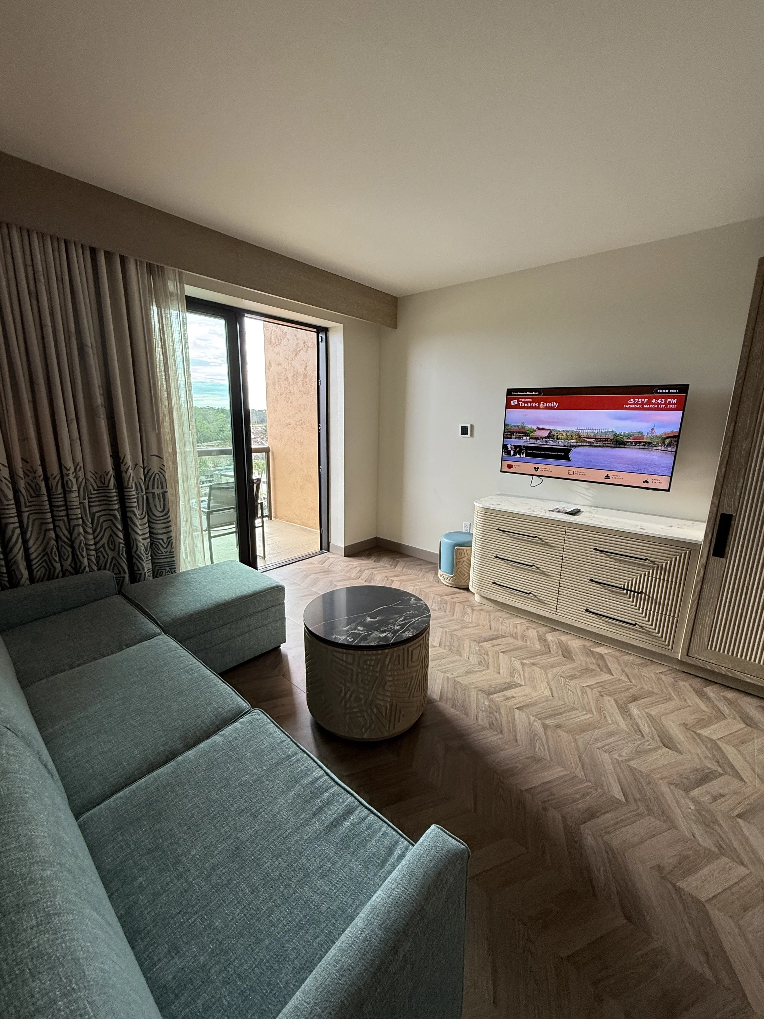 Living room with a sectional sofa, round coffee table, TV mounted on the wall, and sliding glass door leading to a balcony with outdoor furniture.