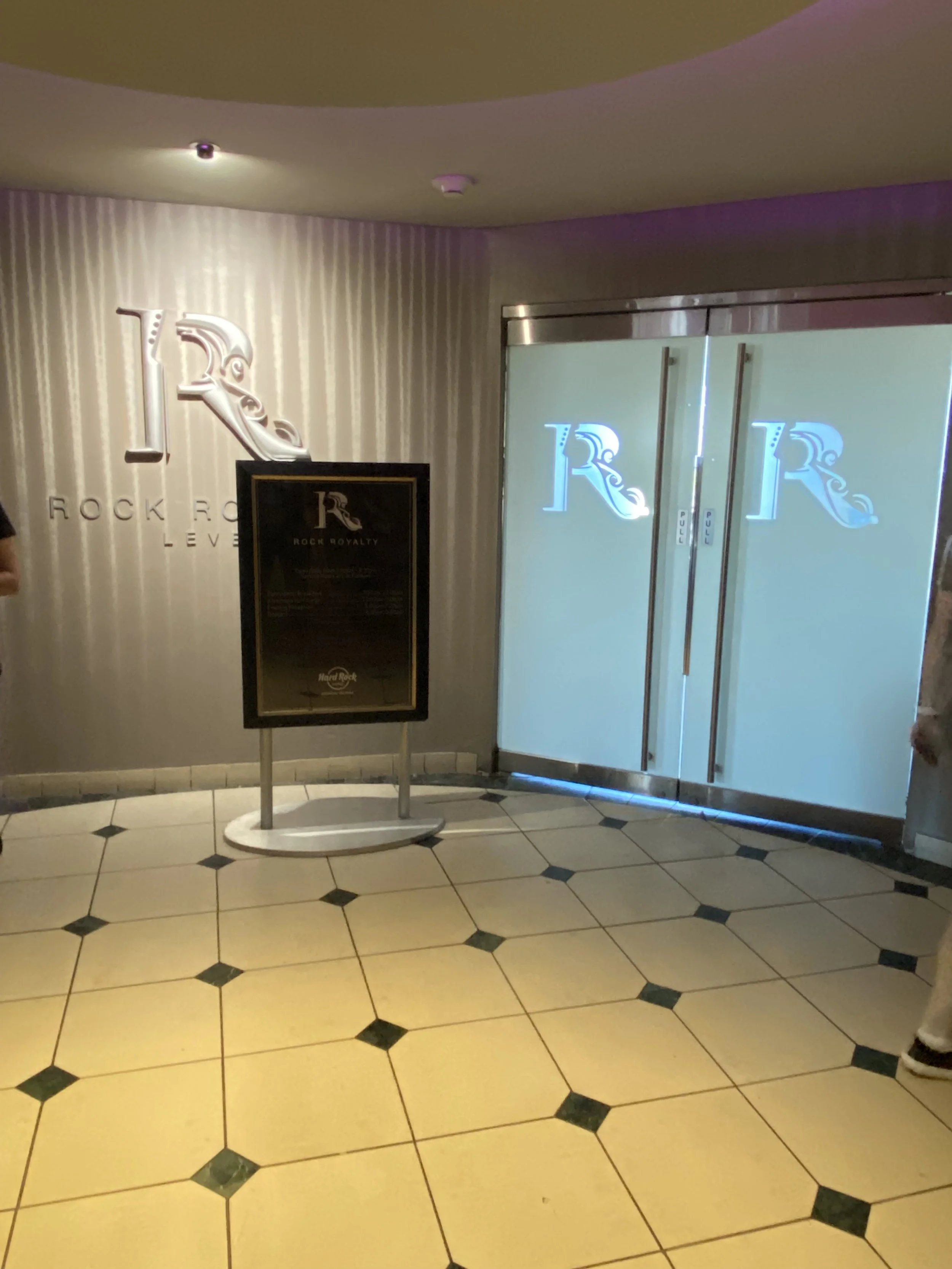 Glass doors with illuminated logos of the Rock Royalty at the entrance of the Hard Rock Cafe, with a signboard stand in front of the wall.
