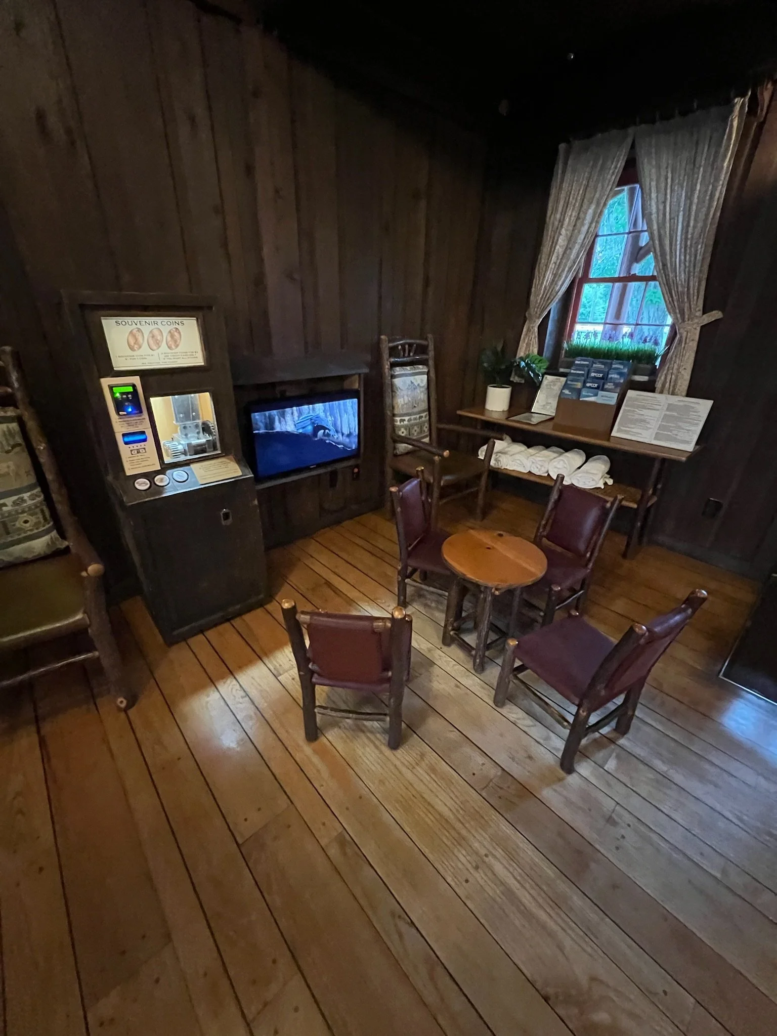 A cozy seating area with three chairs around a small round wooden table, a window with curtains, a vintage coin dispenser, a fireplace, and a table with rolled towels and plants.