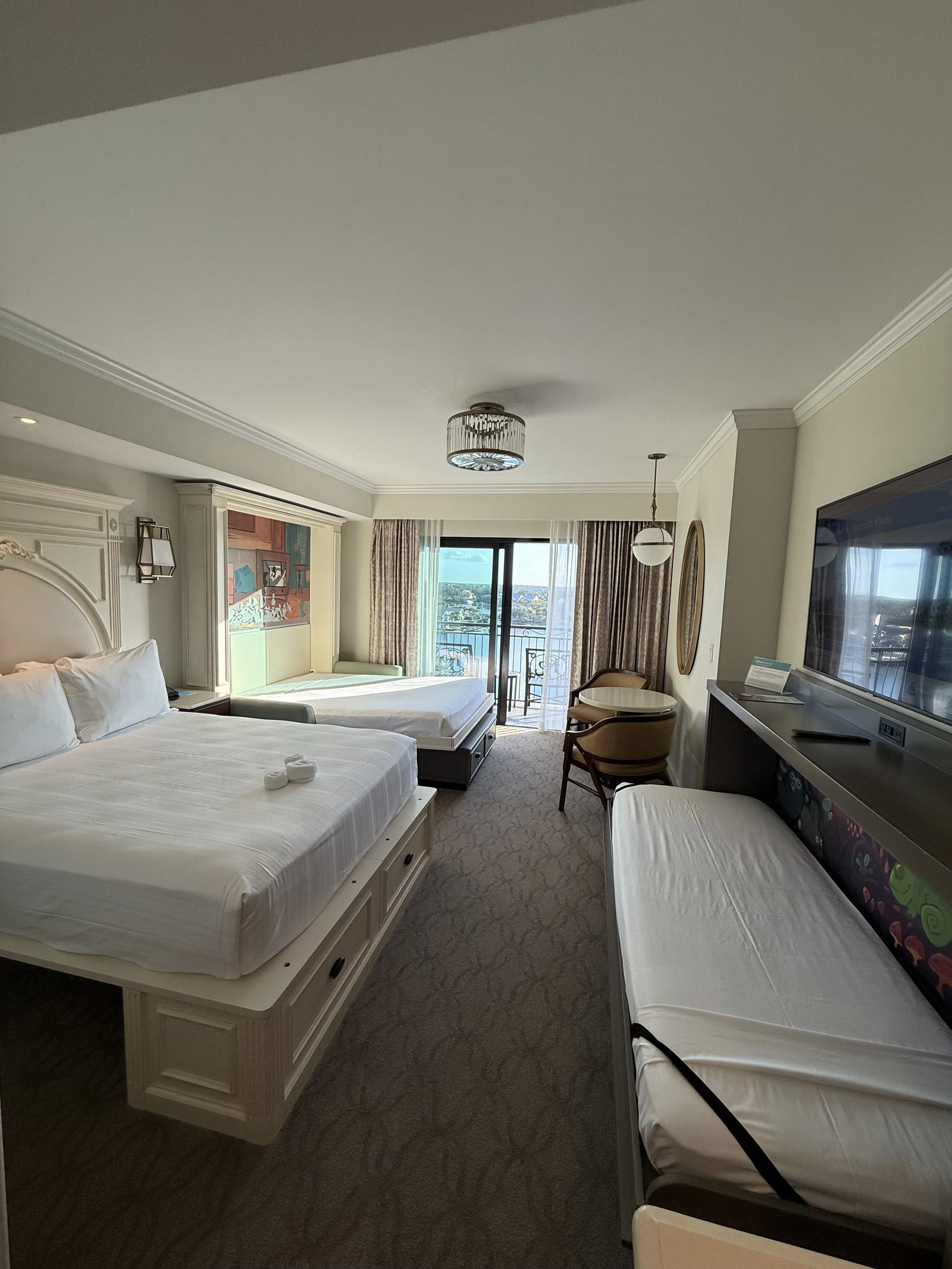 A hotel room with two beds, one larger and one smaller, a balcony with a view, and a sitting area with chairs and a small table.