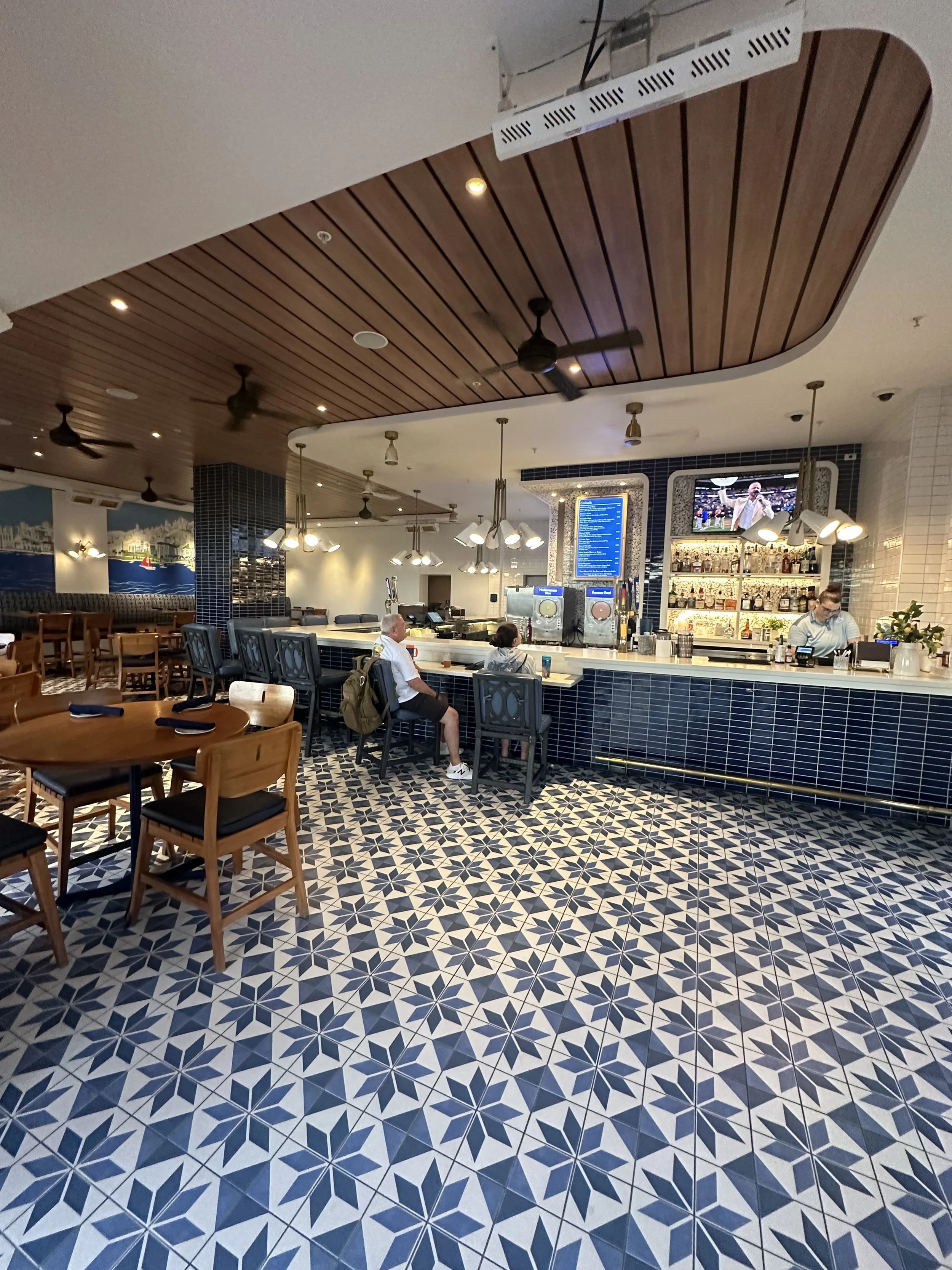 A bar area inside a restaurant with a patterned tile floor, wooden chairs and tables, a bar counter with barstools, and a couple of customers seated.