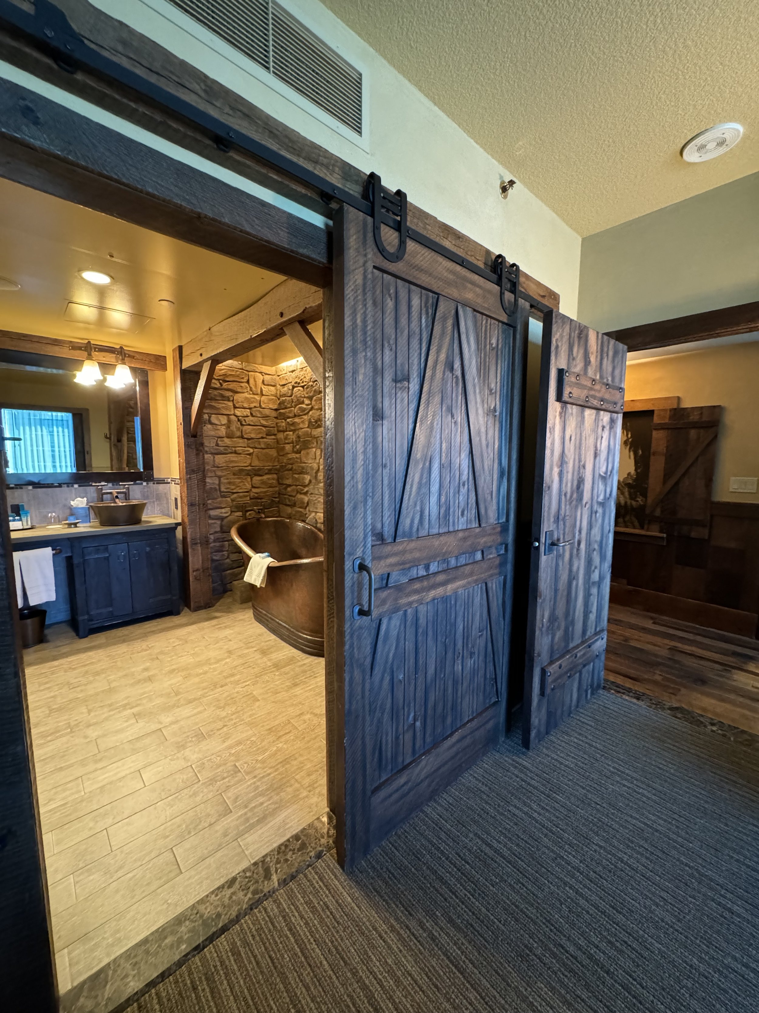 Interior view of a rustic bathroom with a sliding barn door, stone wall, copper bathtub, vanity with vessel sink, and warm lighting.