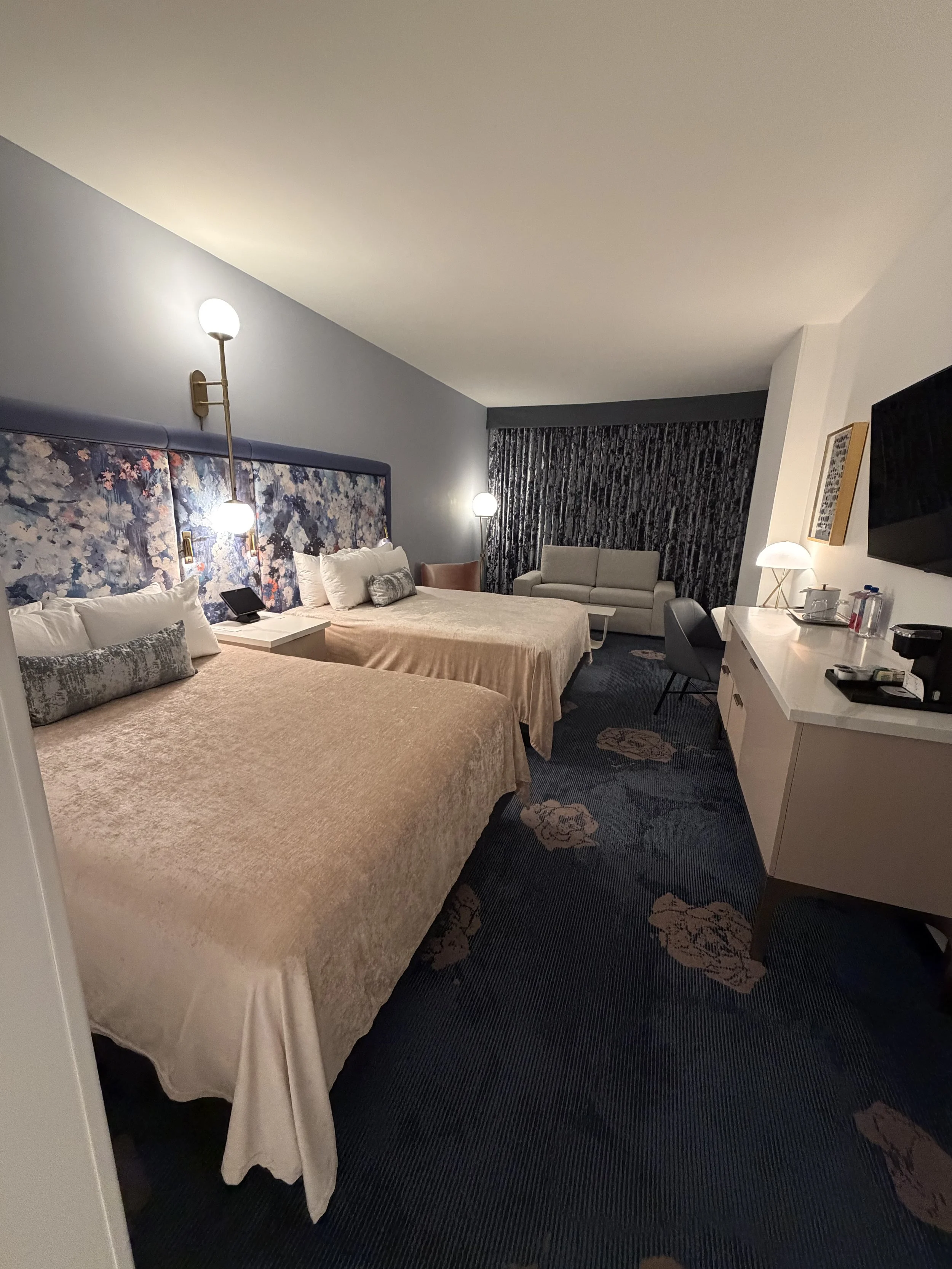 A modern hotel room with two queen beds, floral headboards, bedside lamps, a sitting area with a sofa and chairs, and a dresser with a coffee station and TV.