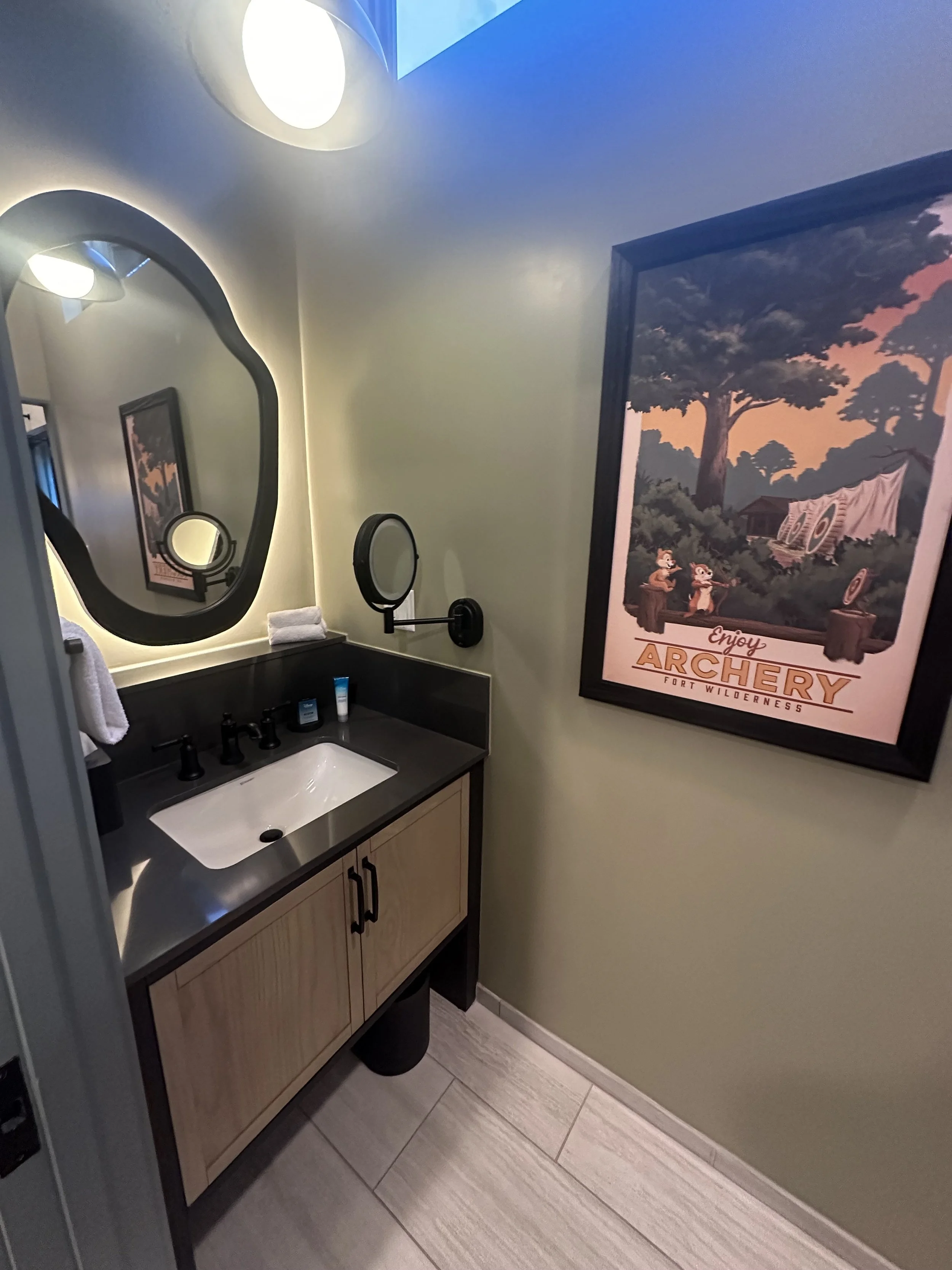 Small bathroom with a black vanity, oval mirror, wall-mounted magnifying mirror, framed archery poster, light green walls, and tiled floor.