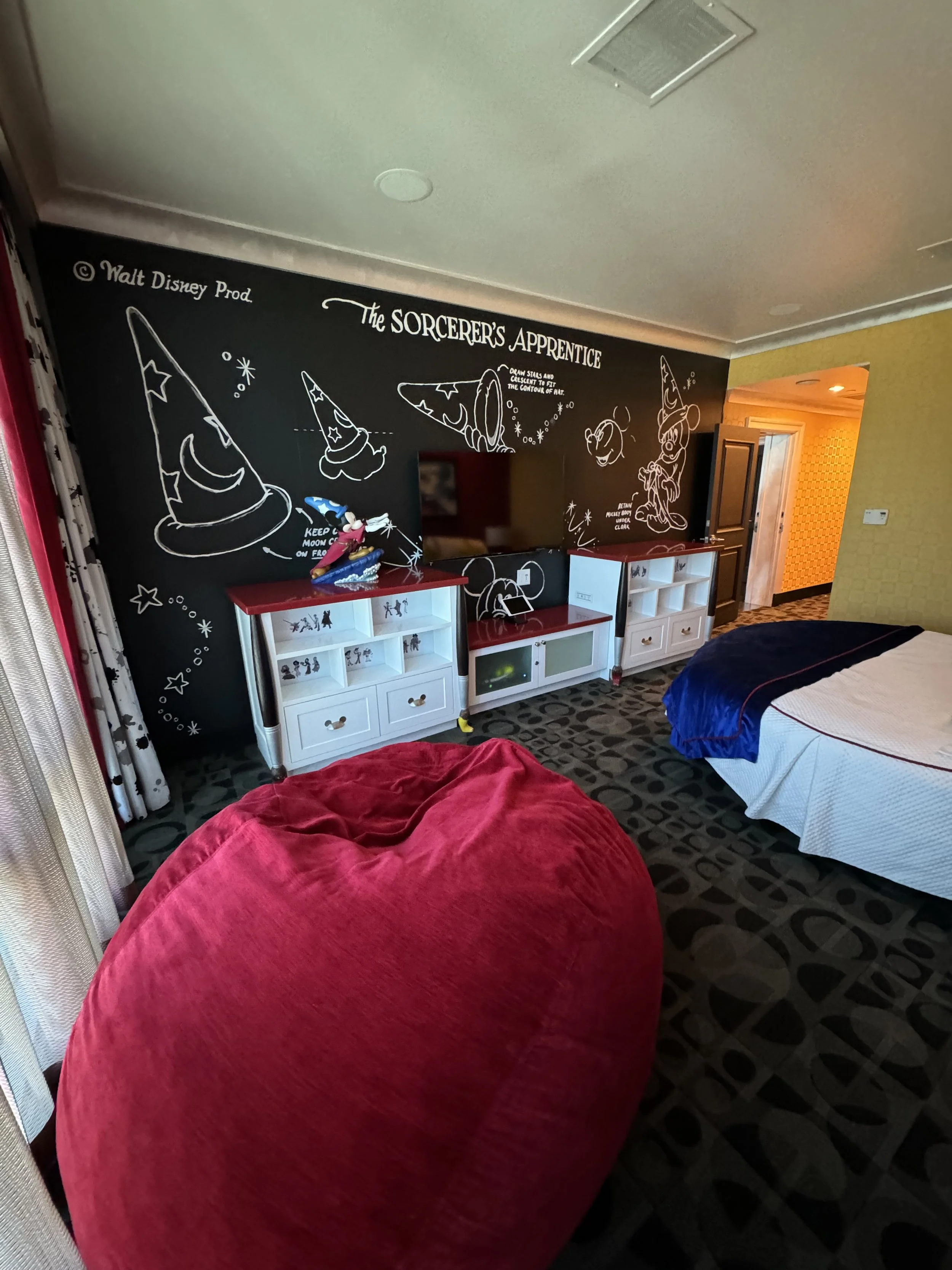 Hotel room with a black chalkboard wall decorated with white illustrations of wizard hats, a broom, and stars, featuring the text 'The Sorcerer's Apprentice.' There are two beds, one with a white bedspread and a blue runner, and a large red beanbag c