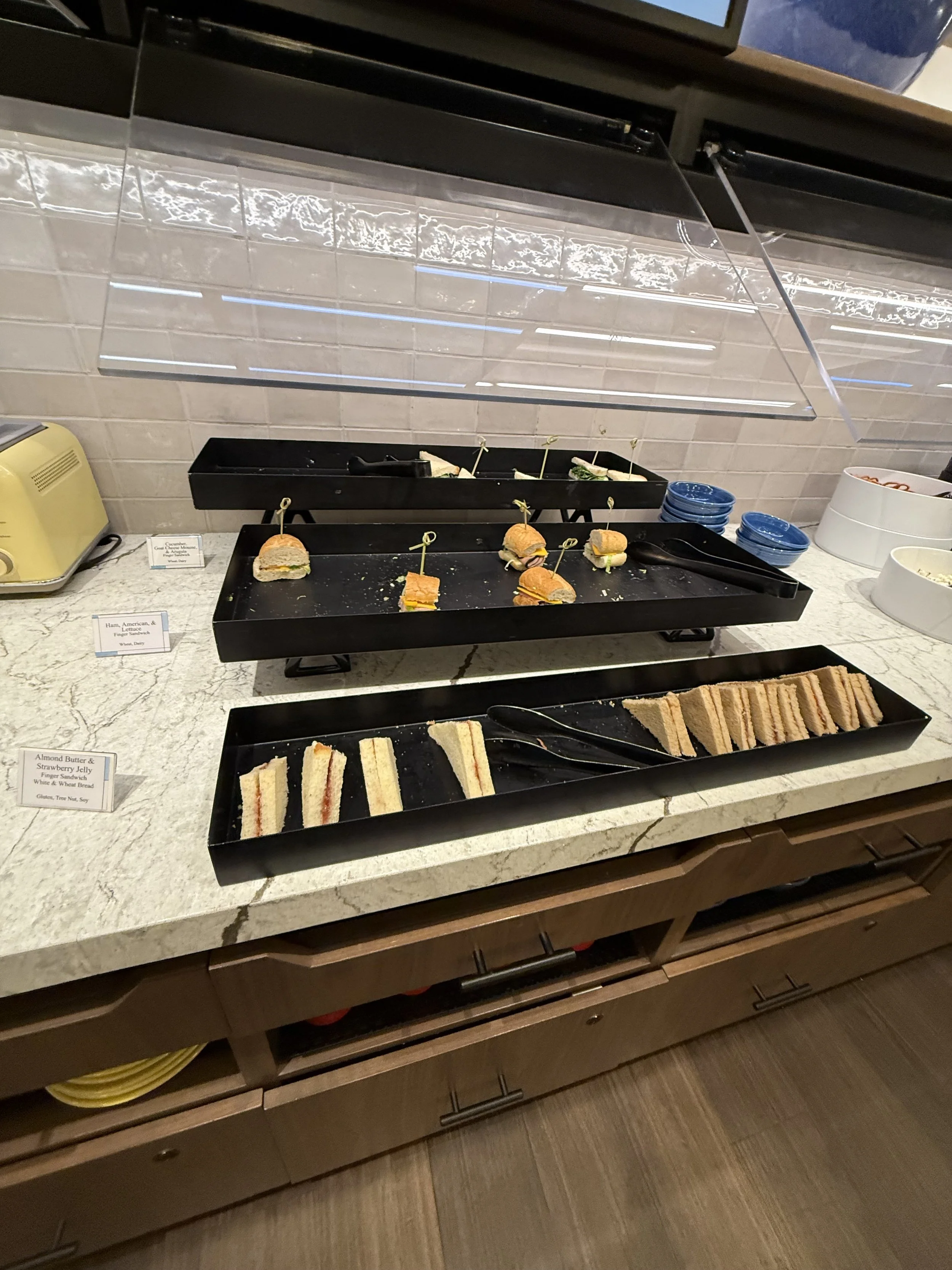 Buffet setup with mini sandwiches, sliders, and cut slices of bread on black trays, with small blue bowls and a white container nearby.