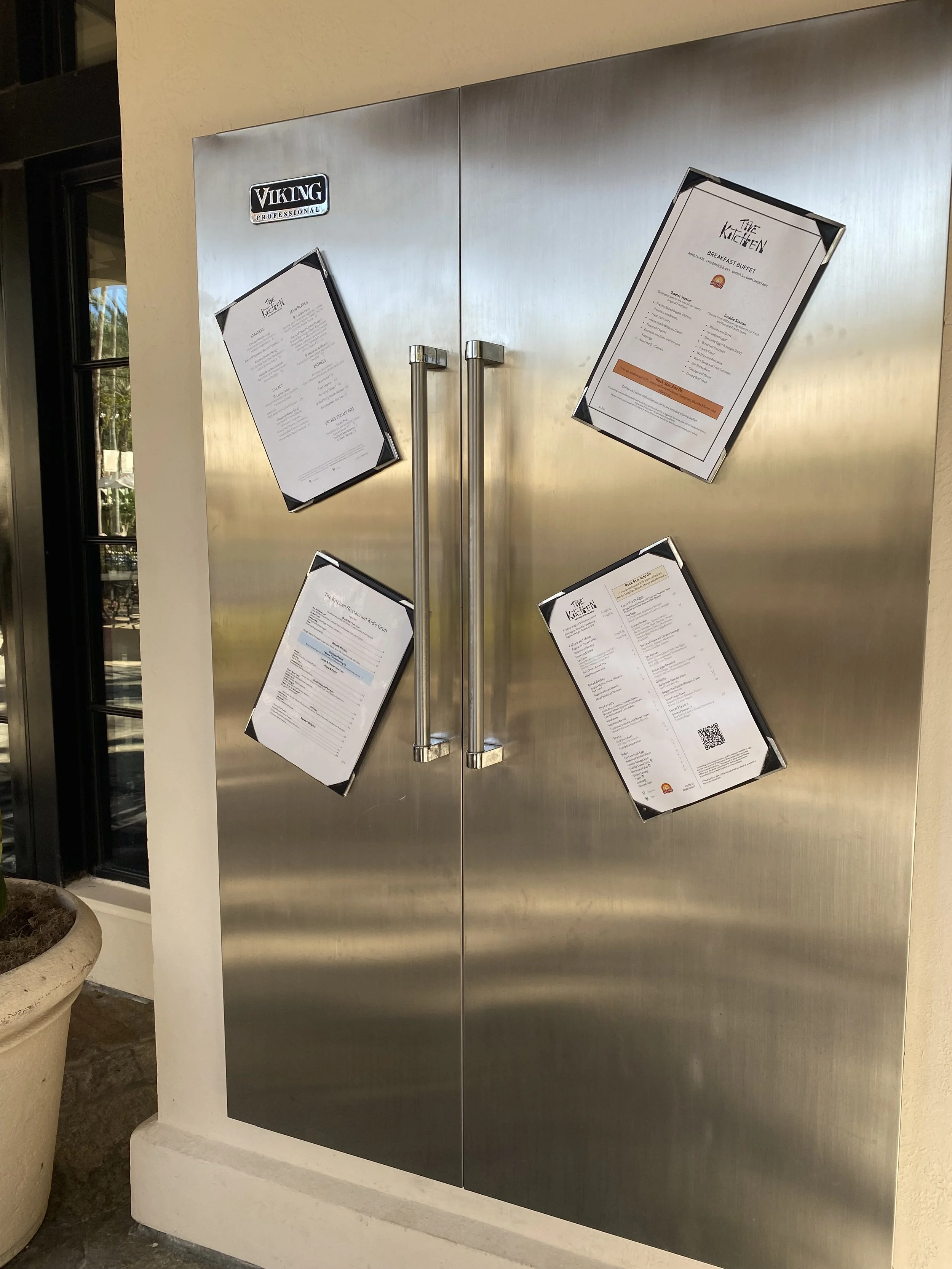 A stainless steel door with four attached restaurant menus displayed at an angle, two on each side, outside a restaurant.