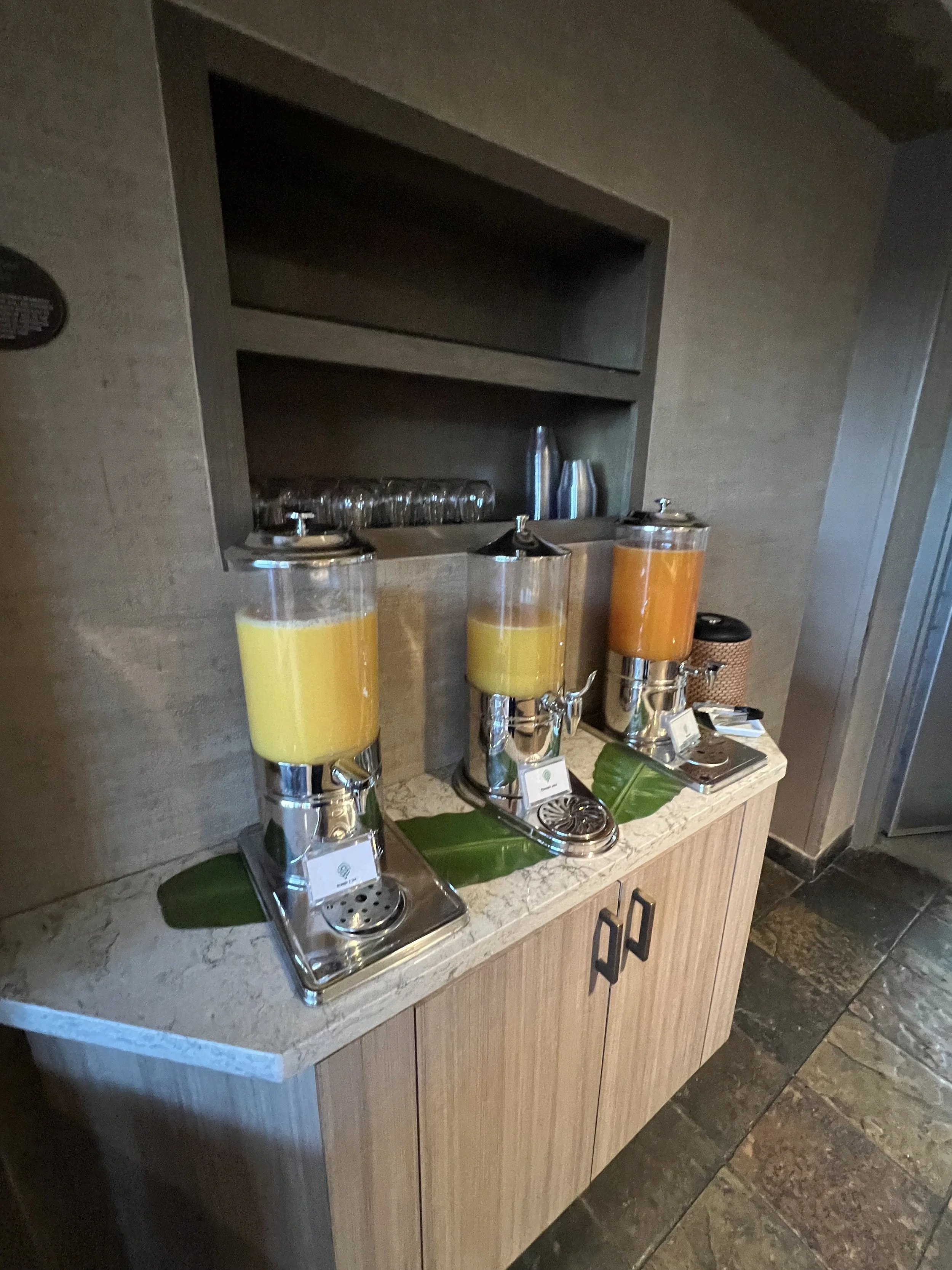 Three glass beverage dispensers filled with yellow, yellow, and orange drinks on a wooden table, with a leaf underneath each dispenser, in a hotel or restaurant setting.