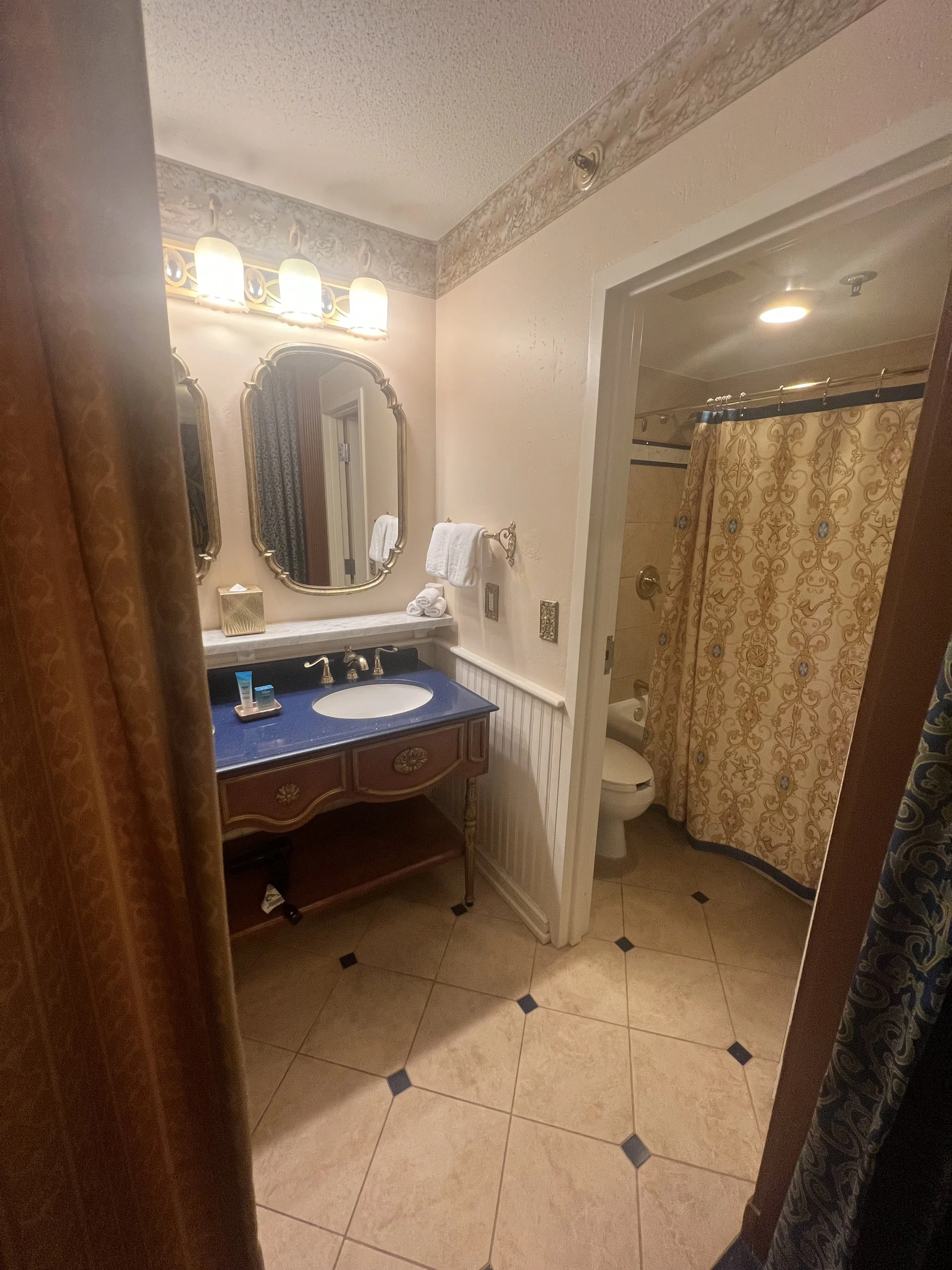 A vintage-style bathroom with a white vanity with a blue countertop, a large mirror, and warm lighting. To the right, a separate toilet area with a decorative shower curtain.