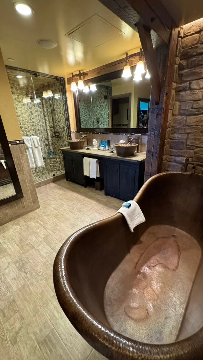 Bathroom with a rustic design featuring a large wooden bathtub, a mirrored vanity with two vessel sinks, a glass-enclosed shower, and warm lighting.