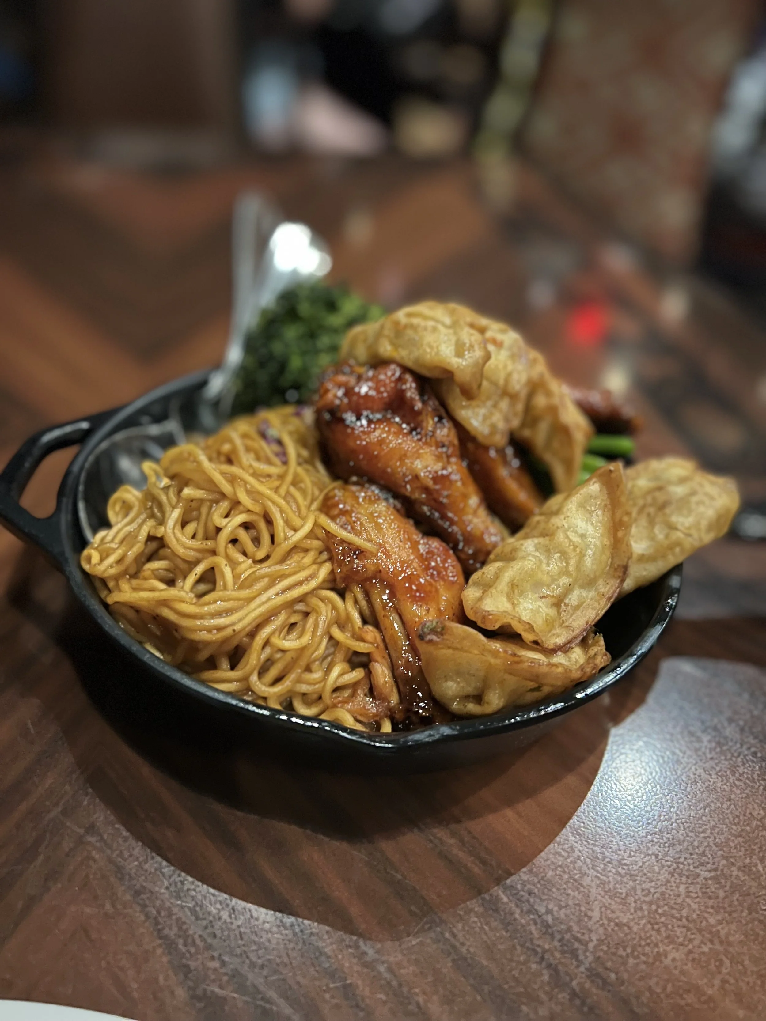 A black bowl filled with Asian cuisine including fried chicken wings, ramen noodles, fried wontons, and steamed broccoli, on a wooden table.