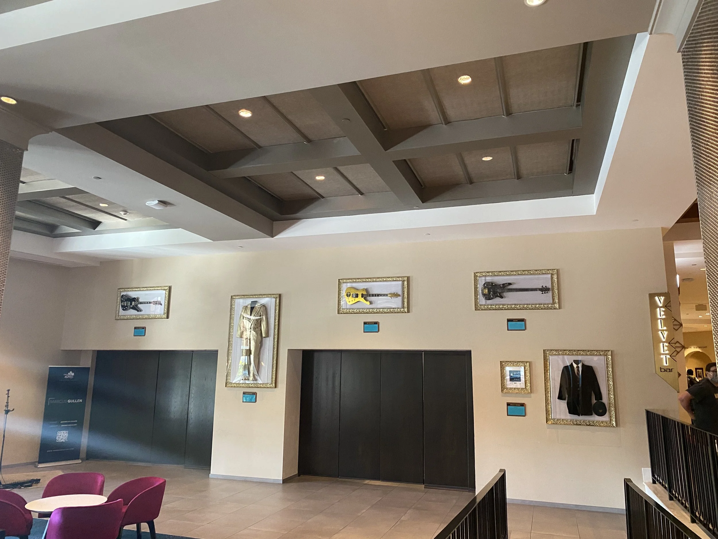 Interior view of a hotel lobby with framed artwork on the wall, including guitars, a suit, and a dress, along with a yellow costume. There is a seating area with pink chairs and a small table, and a sign on the right that says 'VELVET bar.'