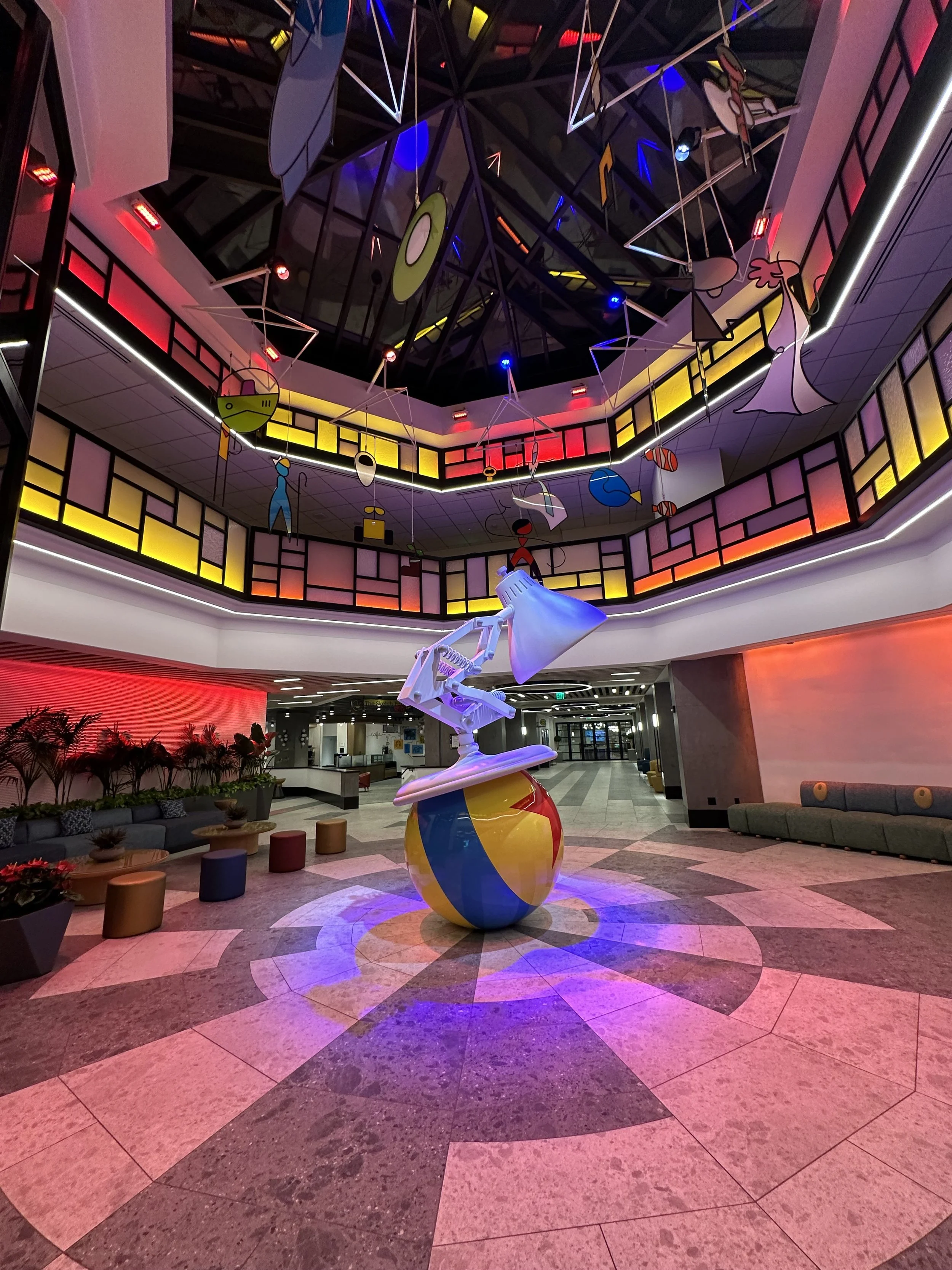 Interior of a modern lobby with colorful lighting, a large striped beach ball sculpture, and abstract hanging art pieces on the ceiling. The space features a seating area with green couches, small round stools, and decorative plants.
