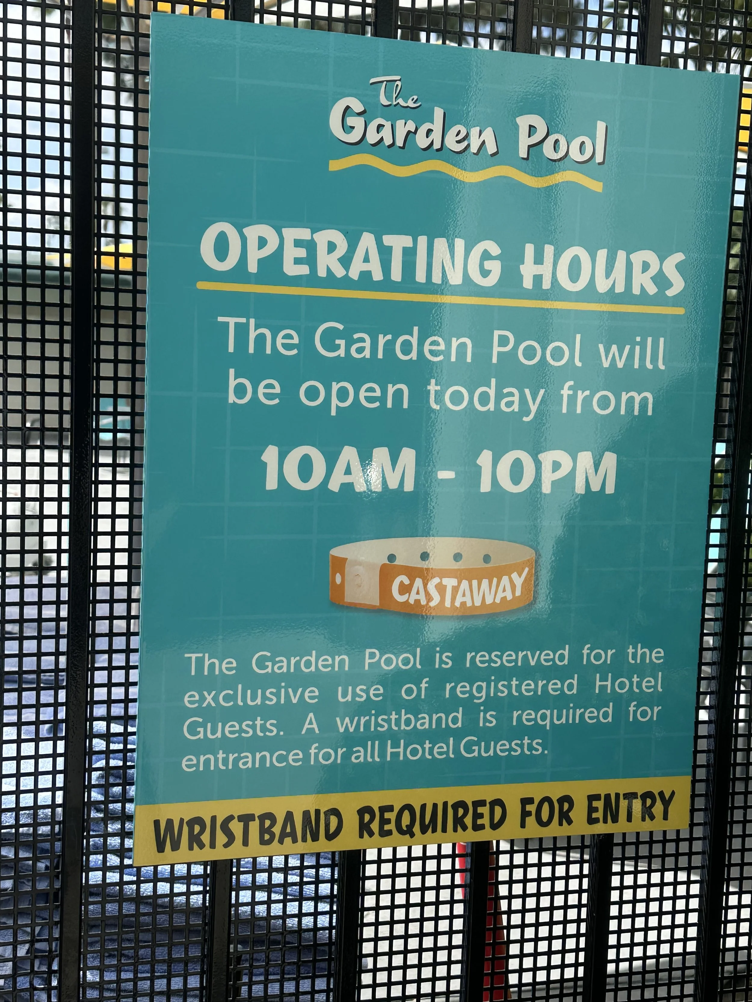 Signboard displaying the operating hours of the garden pool, which is from 10AM to 10PM, and mentioning that a wristband is required for entry, reserved for hotel guests, with a diagram of a wristband labeled "Castaway."