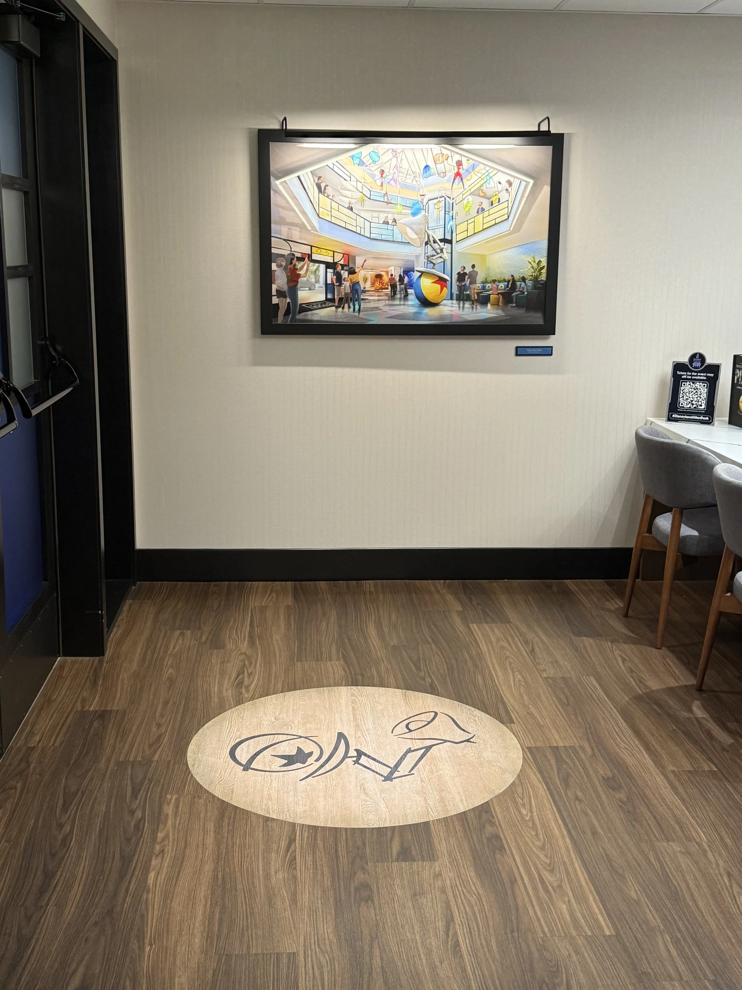 Interior of a building, featuring a framed photo on the wall, a wood floor with a logo created in the floor design, and chairs along a wall with a table that has a QR code and a payment device.