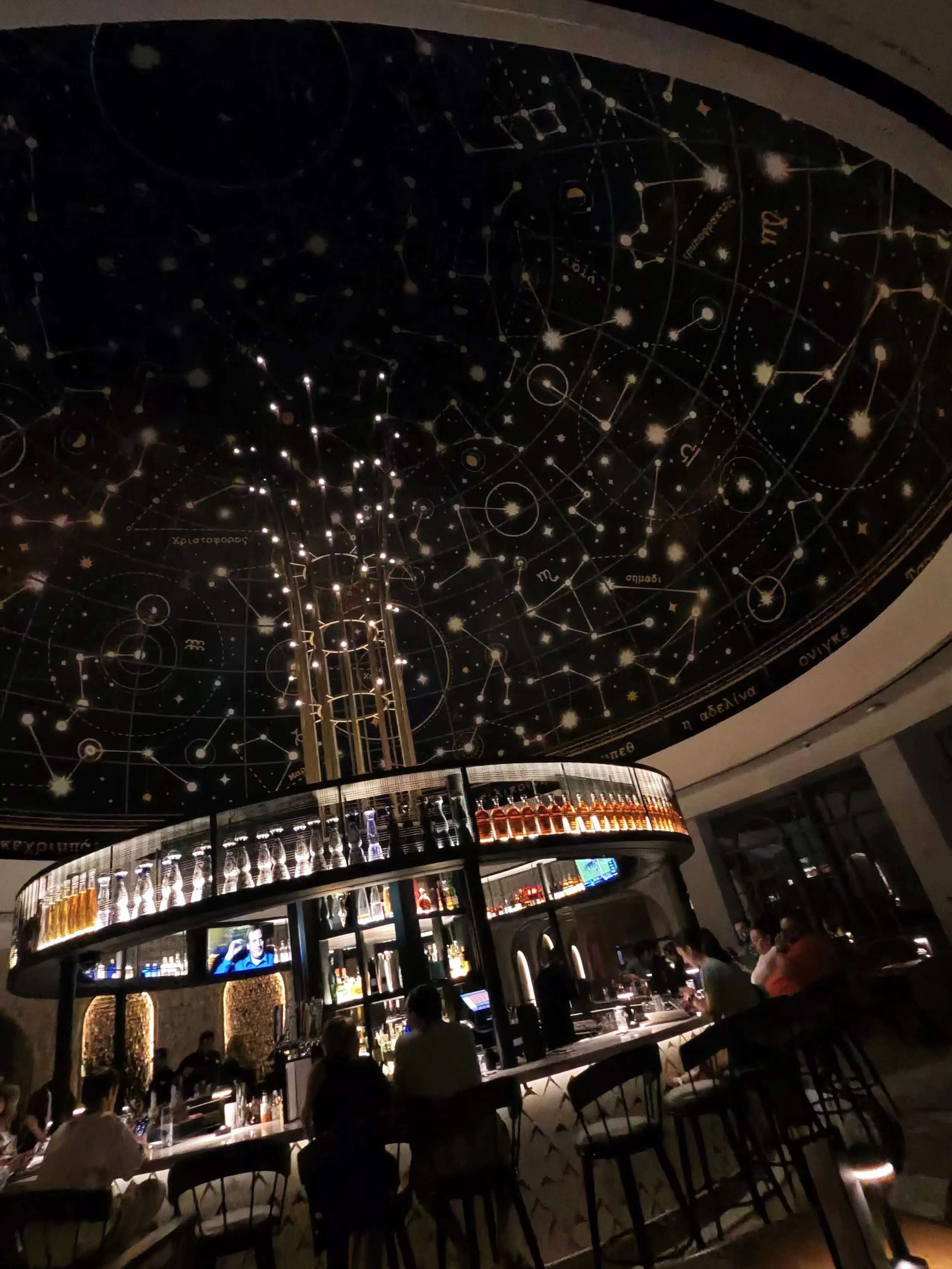 Interior of a bar with a circular counter, illuminated bottles, and a large ceiling mural of a starry night sky with constellations.