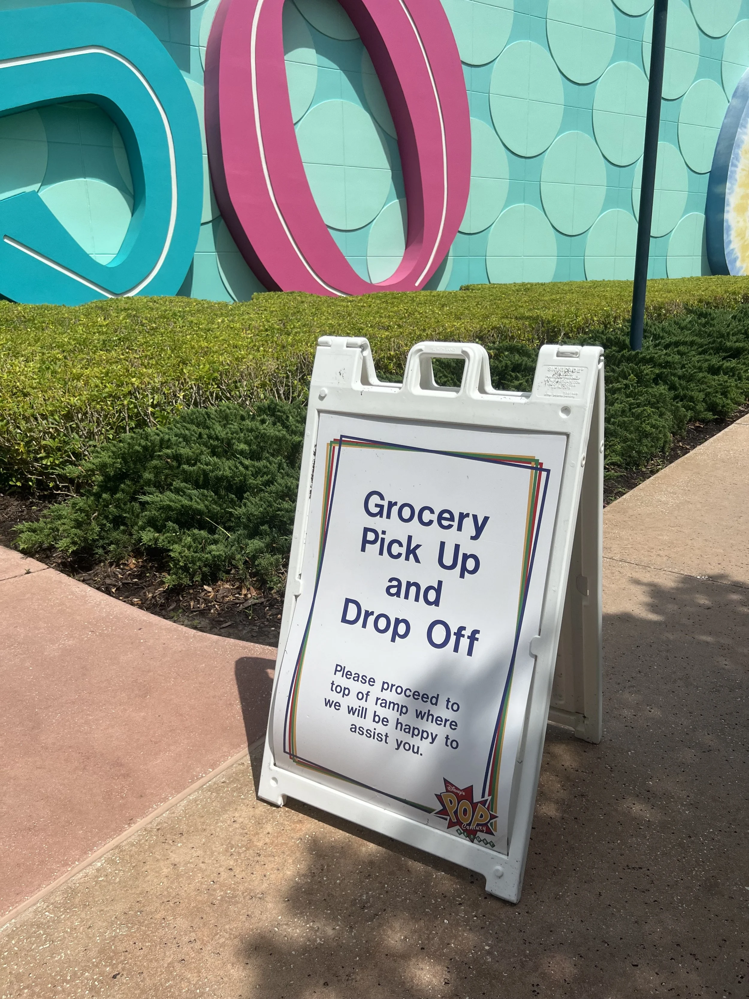 A white signboard with blue text that reads 'Grocery Pick Up and Drop Off. Please proceed to top of ramp where we will be happy to assist you.' The signboard also has colorful borders and a small logo at the bottom right corner. It is placed on a pav