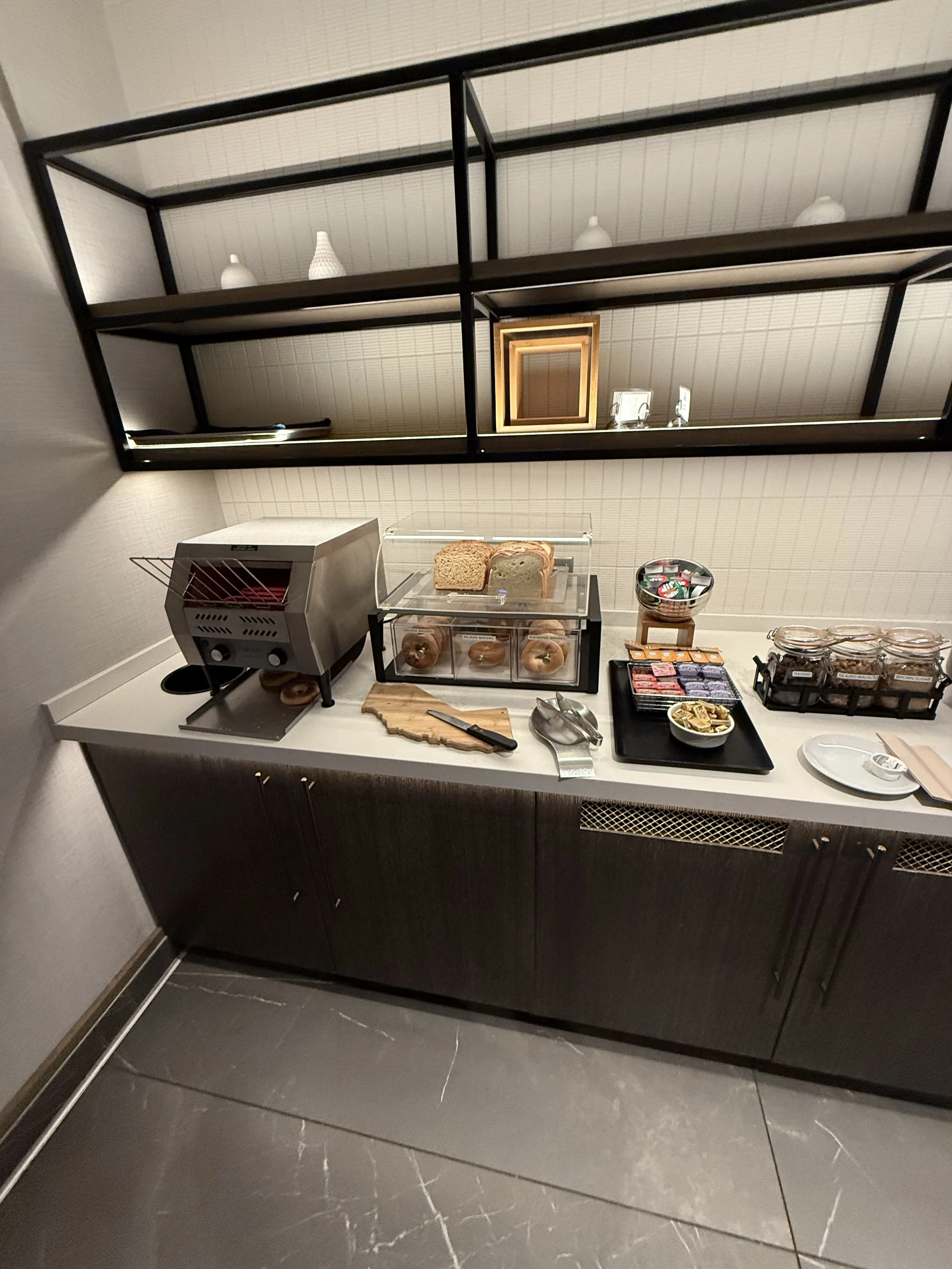 Breakfast buffet setup with bread, pastries, and toppings, on a white countertop with black cabinets and open shelving above.