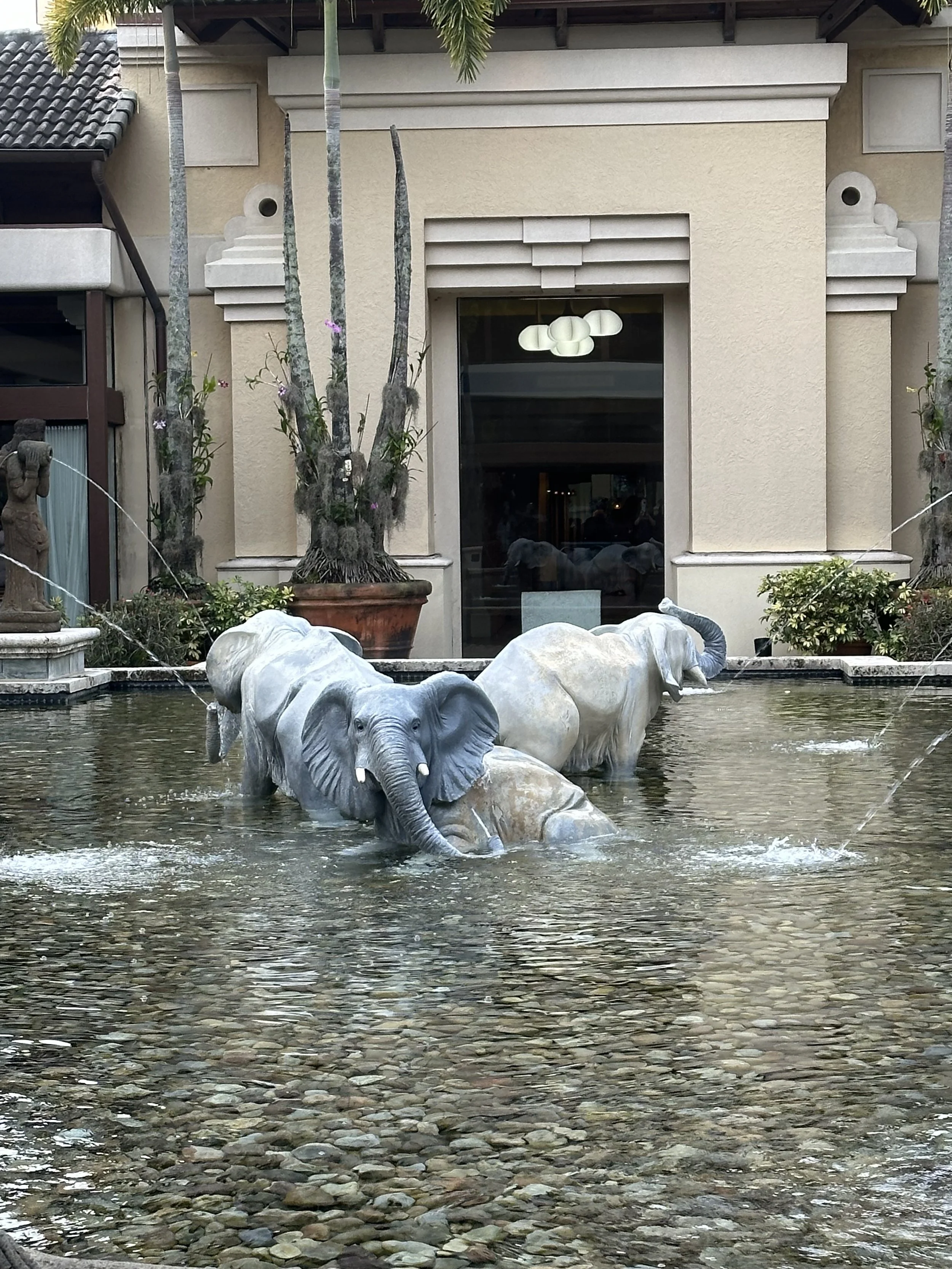 Decorative elephant statues in a fountain with water, located in front of a beige building with windows and palm trees.