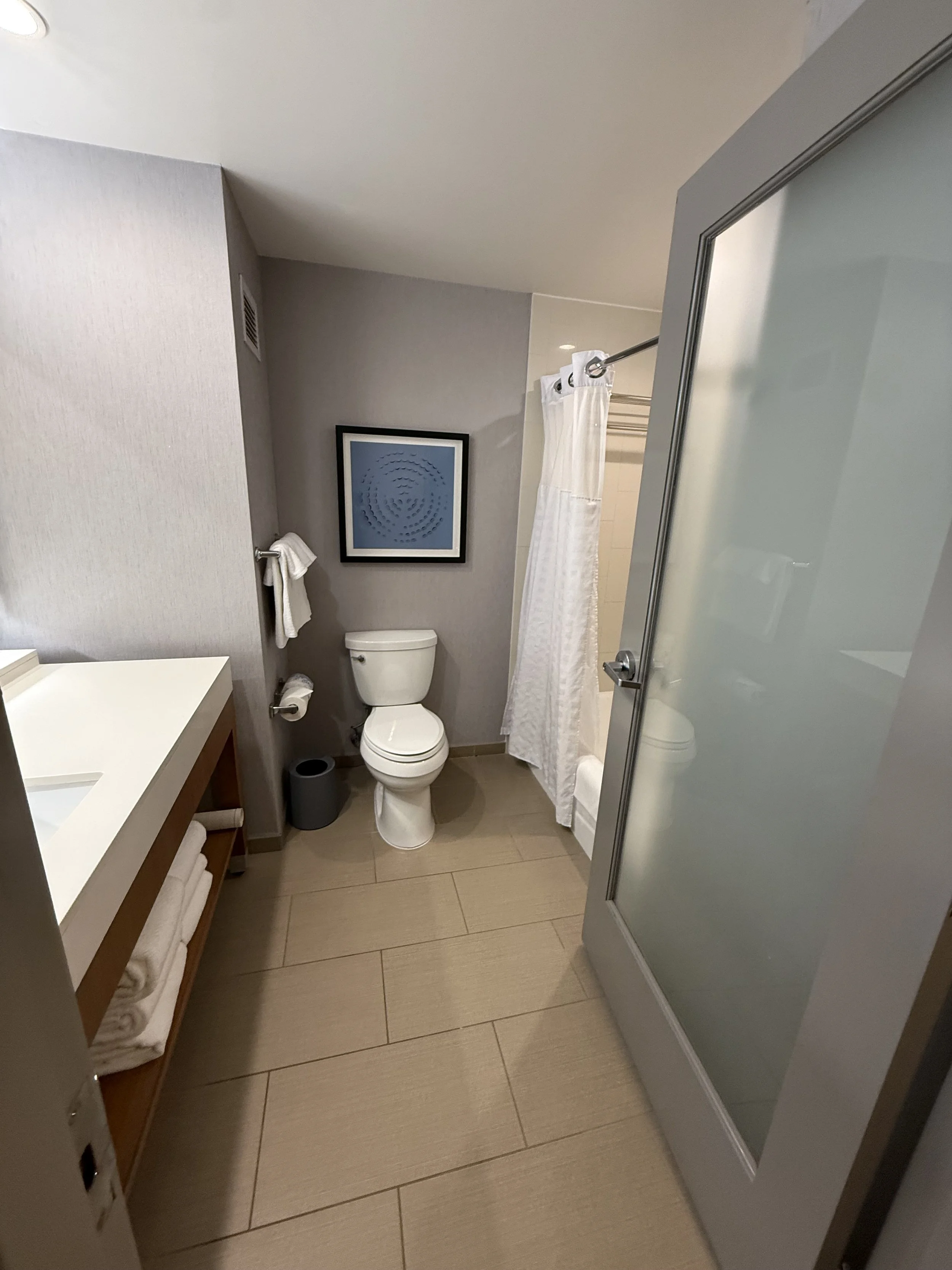 A hotel bathroom with a toilet, a vanity with folded towels, a shower with a frosted door, and a framed abstract art piece on the wall.