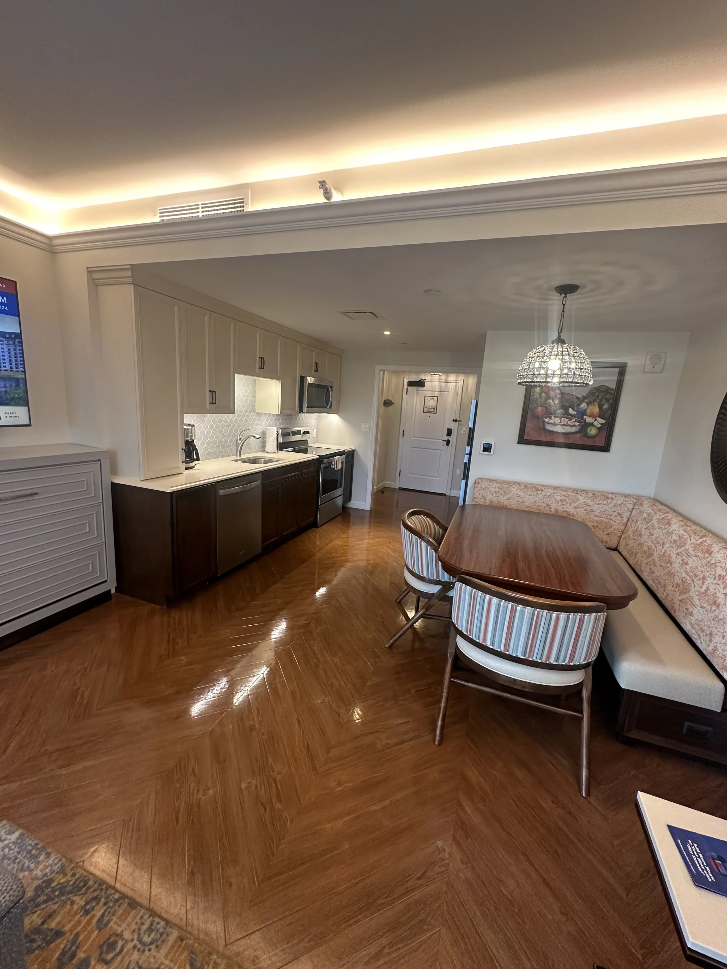 Interior of a hotel room suite showing a kitchenette with cabinets, a dining area with a wooden table, striped chairs, a bench with floral upholstery, and a hanging chandelier, with a door in the background.
