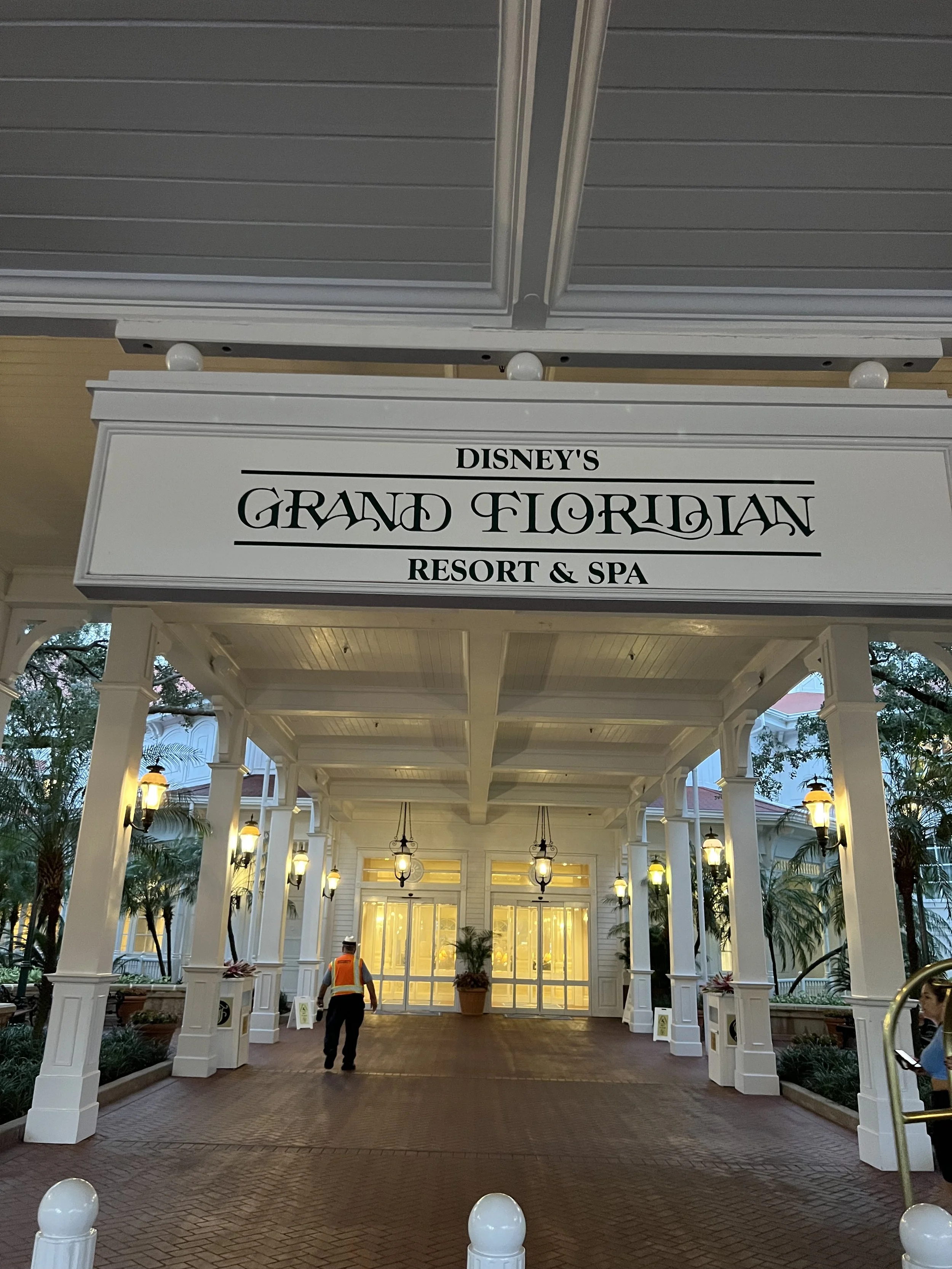 Entrance to Disney's Grand Floridian Resort & Spa with a white sign, decorative pillars, warm lighting, and a person walking in a high-visibility vest.