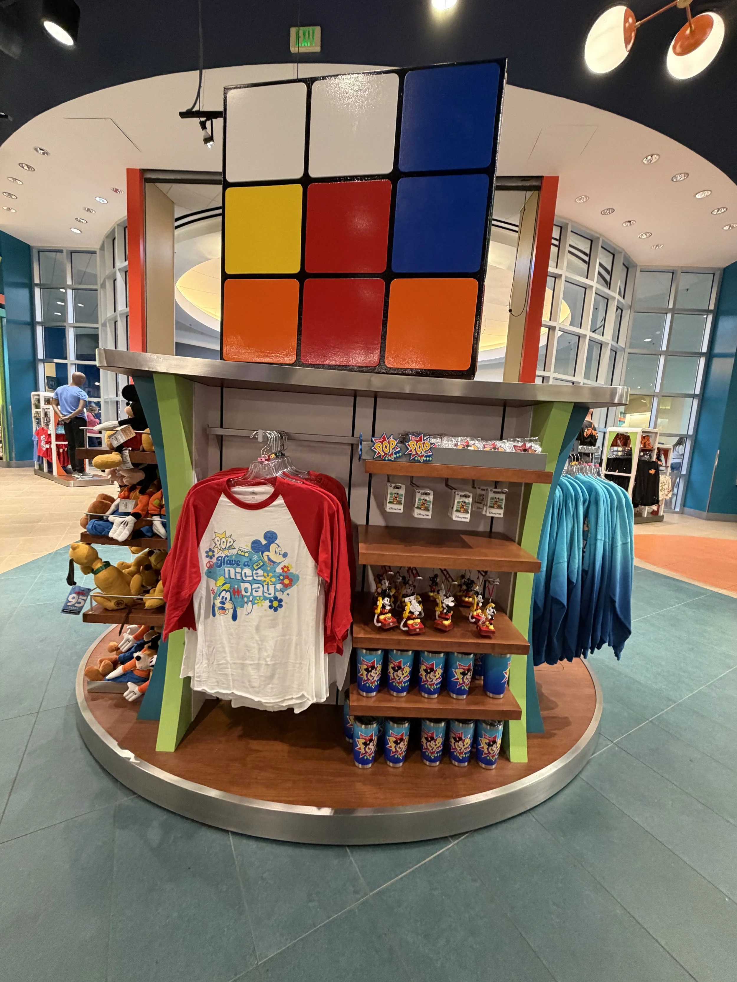 A merchandise display with a large Rubik's Cube on top, and Disney-themed clothing and toys centered around Mickey Mouse, located inside a colorful shopping area.