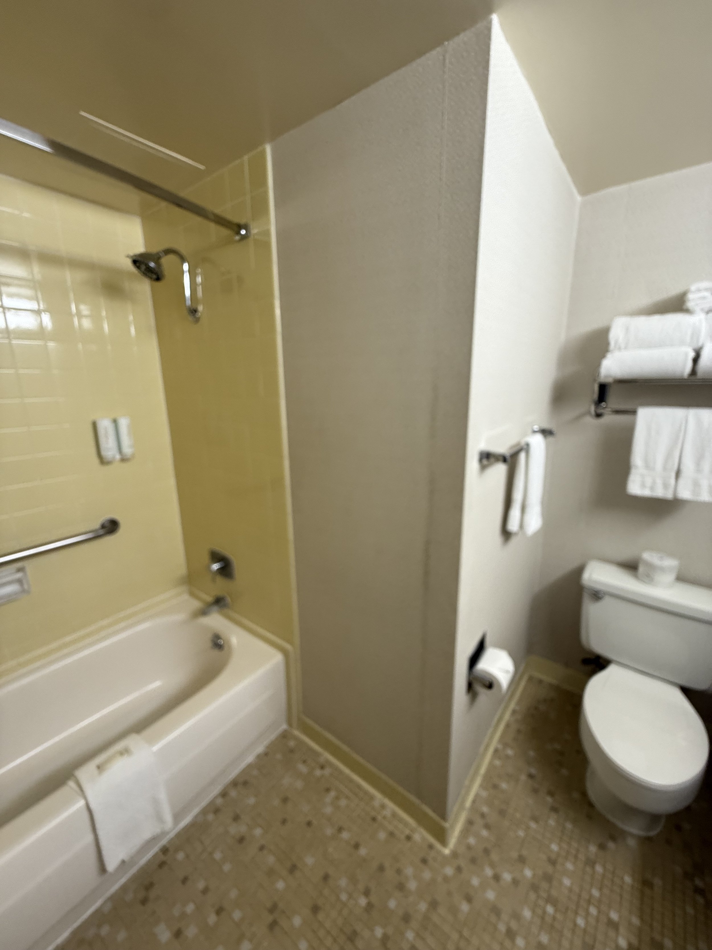 Small hotel bathroom with a bathtub, shower, and toilet, with multiple white towels on shelves and racks.