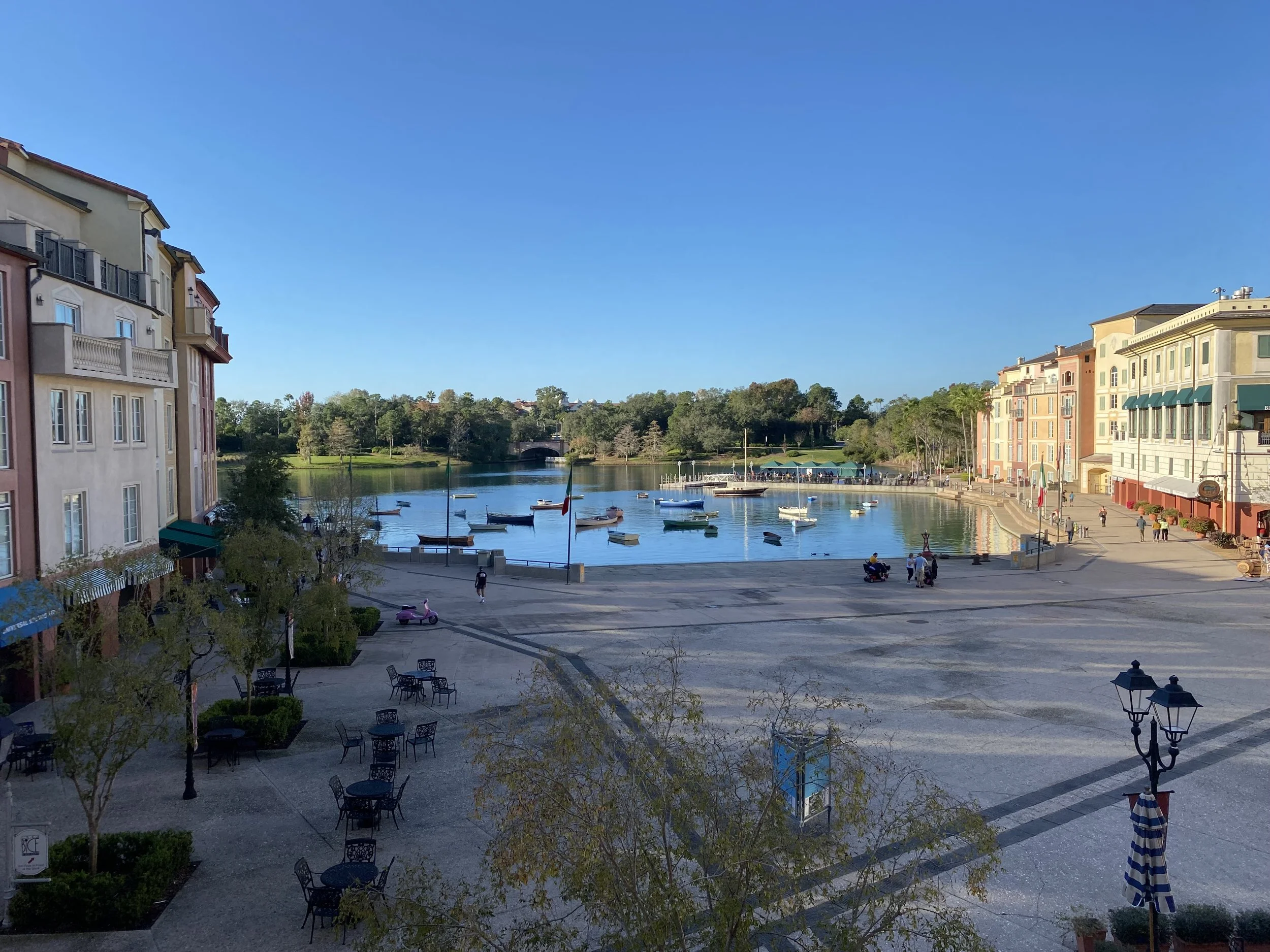 View of a waterfront shopping area with colorful buildings, boats docked in the water, and people walking along the promenade on a sunny day.