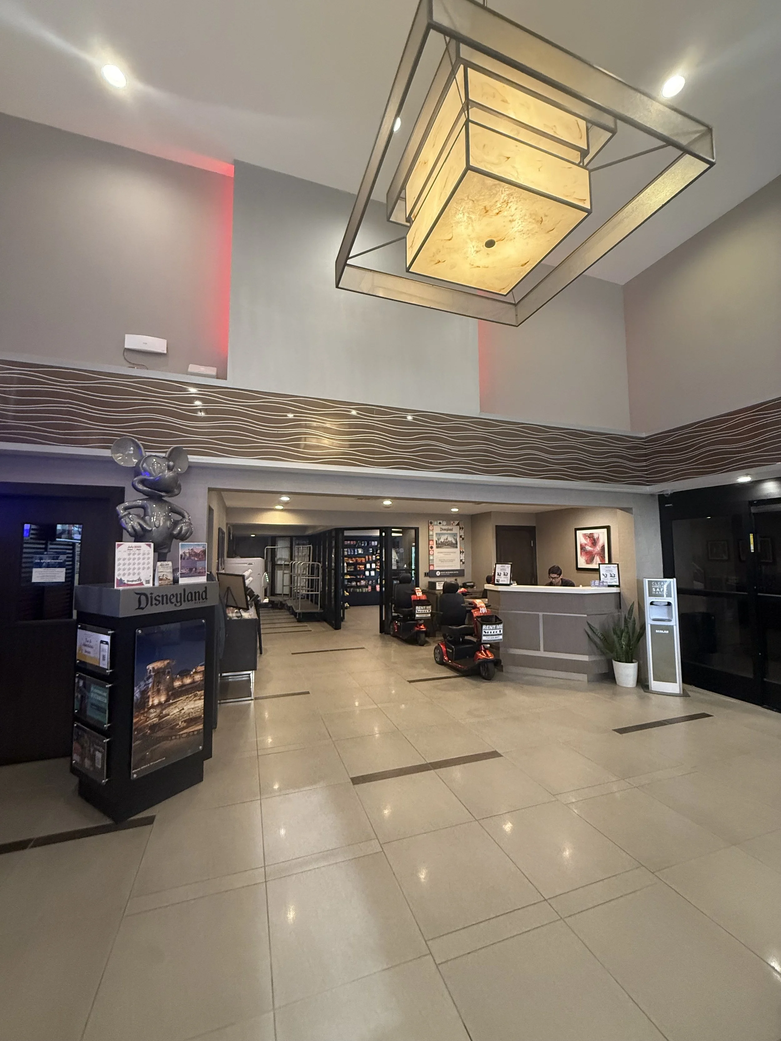 Hotel lobby with a front desk, seating area, and kids' ride-on cars. Modern lighting and decor, with a flower painting on the wall.