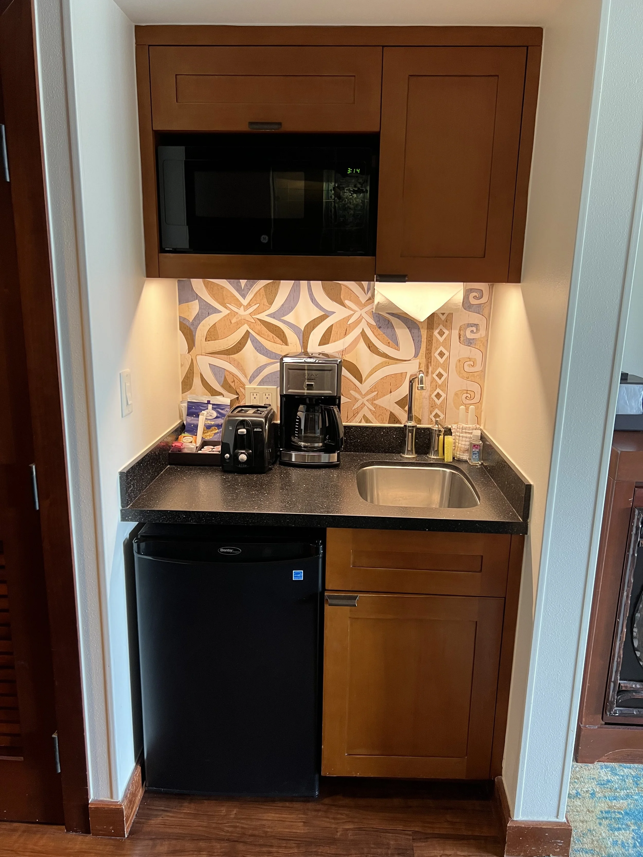 A kitchenette with wooden cabinets, a black microwave, a coffee maker, a toaster, and a mini fridge. The backsplash has a decorative tile pattern, and there is a small sink with a yellow sponge and soap behind the sink.