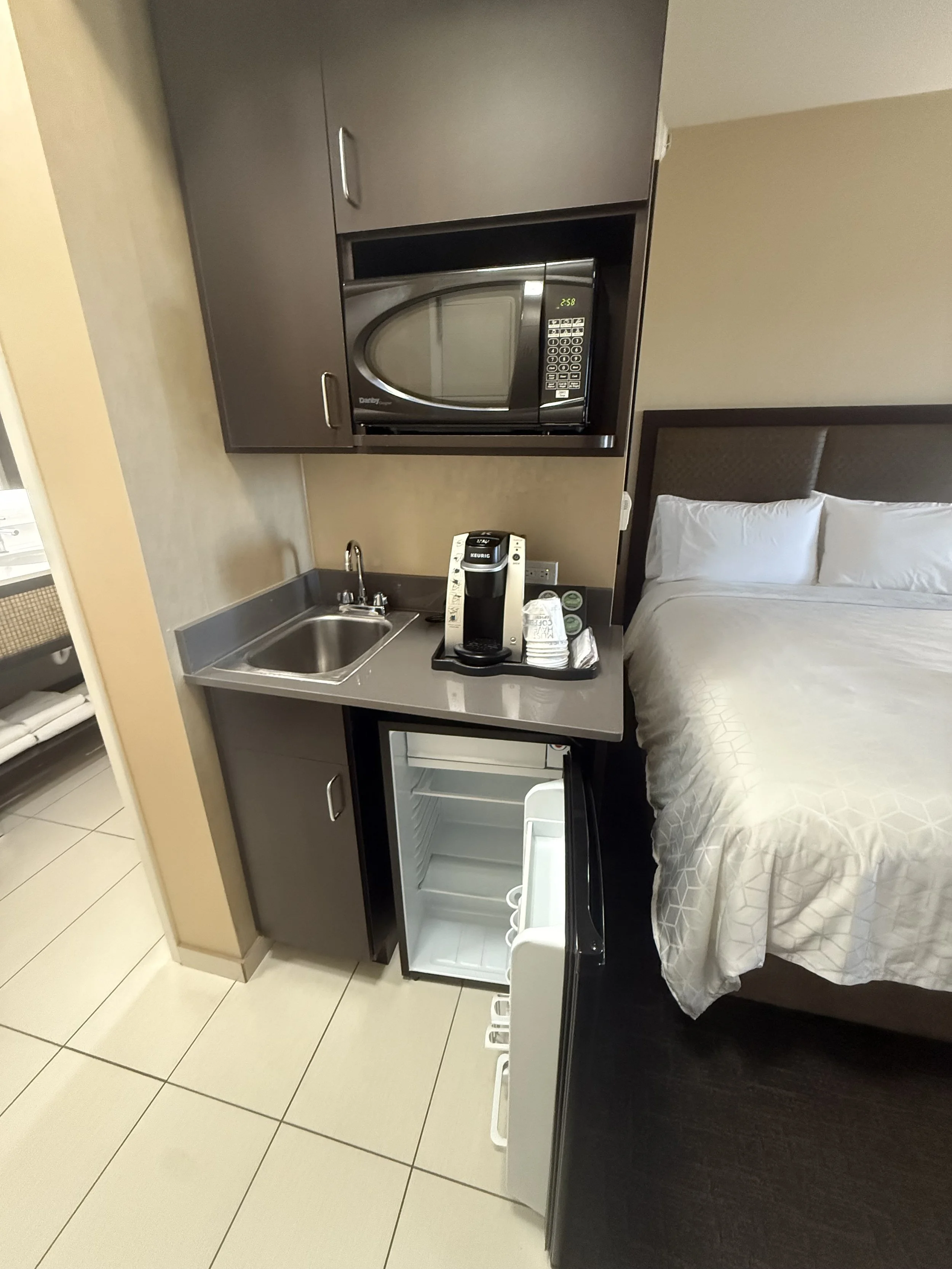 Hotel room kitchenette with microwave, mini fridge, coffee maker, sink, and bed.