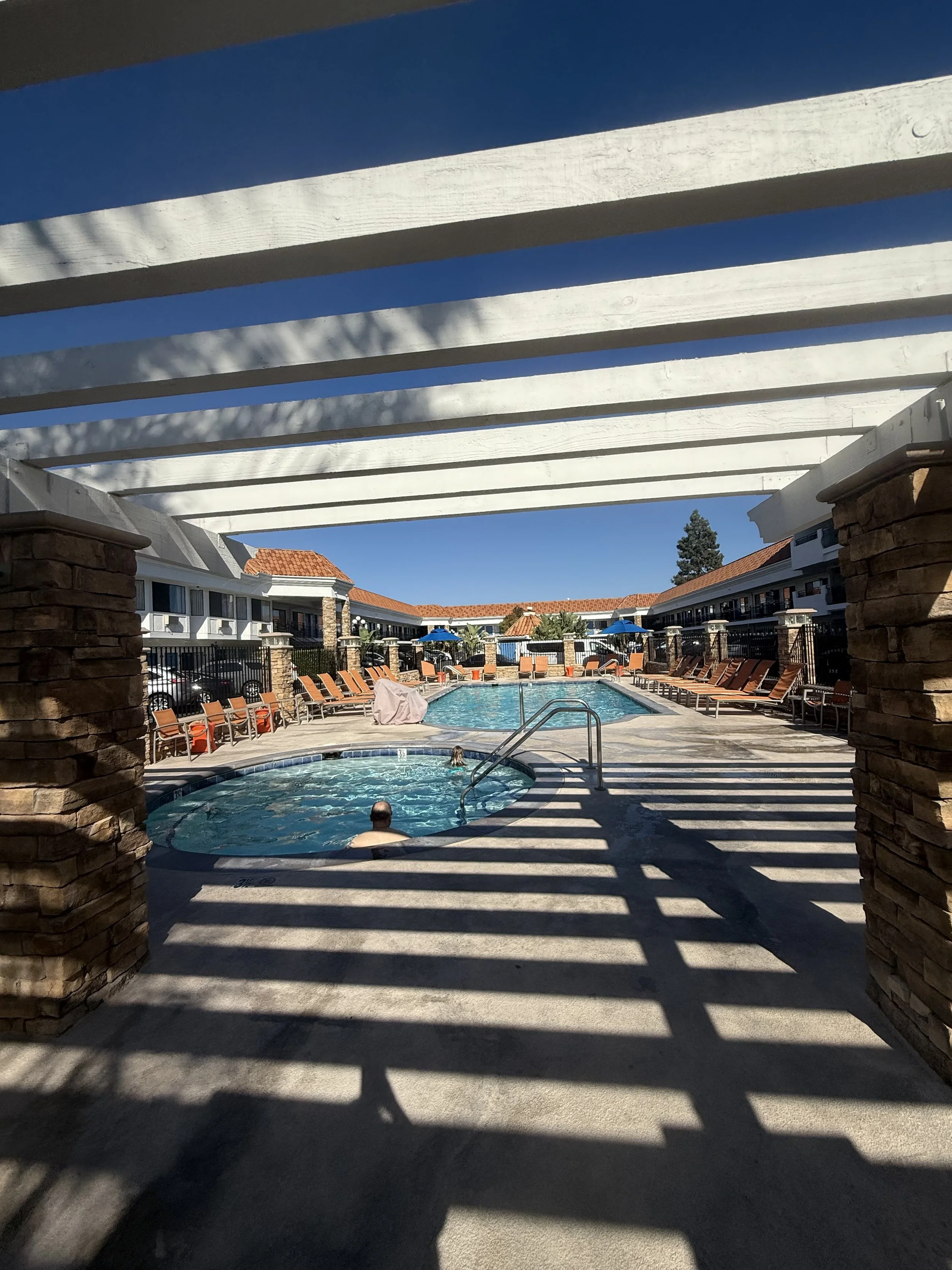 View of an outdoor swimming pool area in a hotel courtyard, with lounge chairs, umbrellas, and a hot tub, seen through white overhead beams casting shadows.