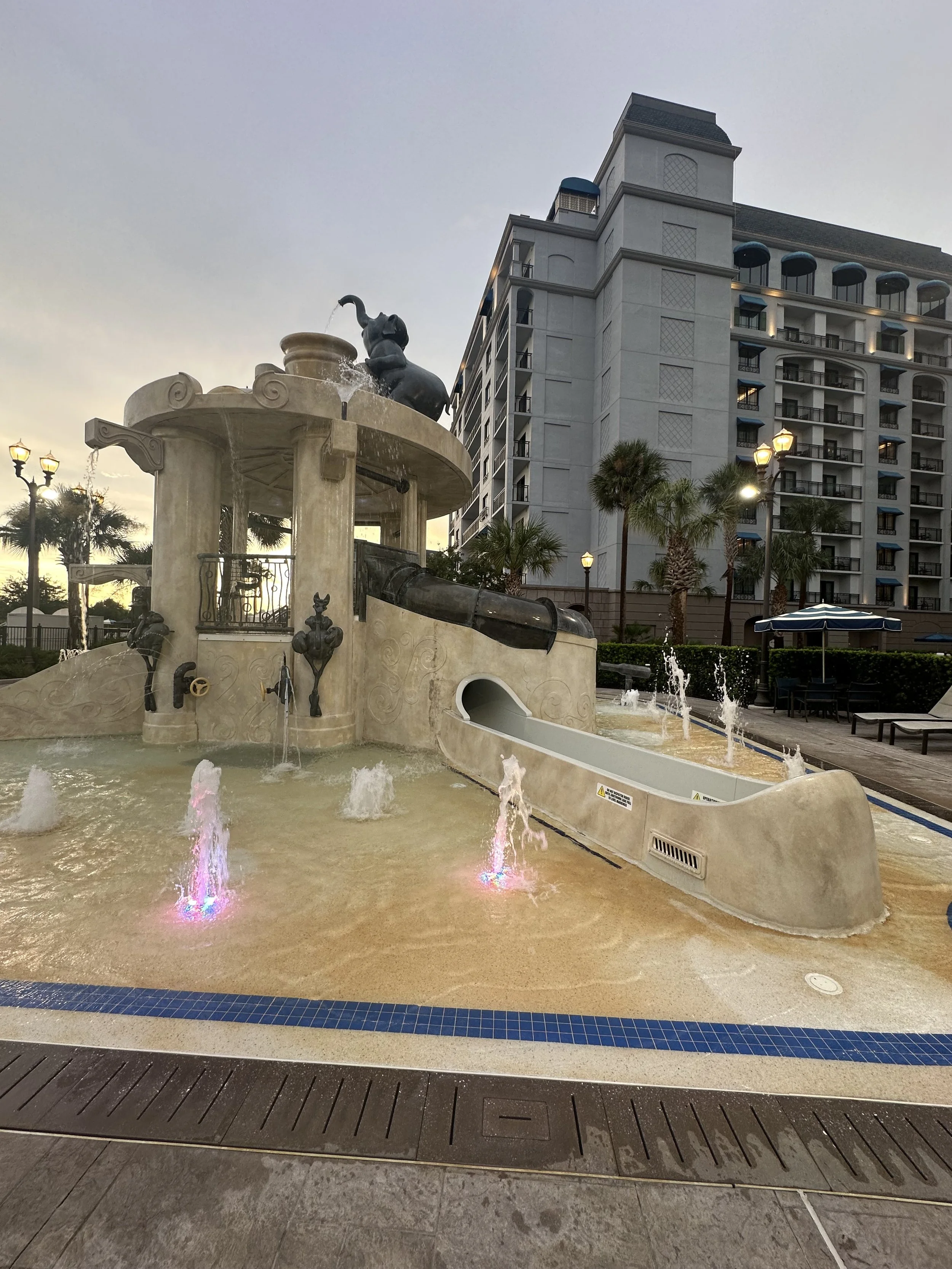 A decorative water fountain with a small slide, featuring a statue of an elephant on top, located outdoors near a modern apartment complex and palm trees at sunset.