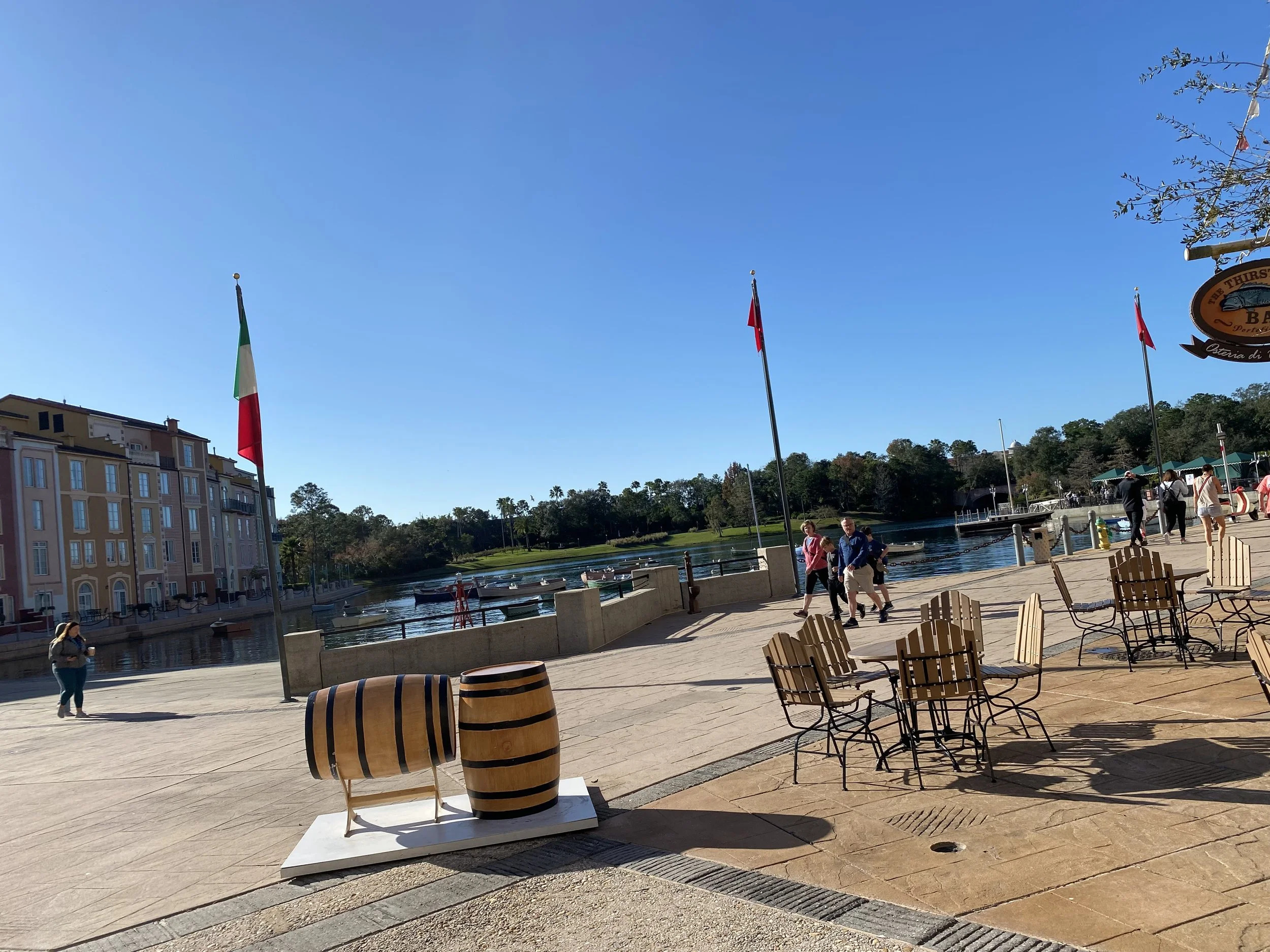 A waterfront scene with outdoor seating, boats on the water, and flags, including the Italian flag, flying on flagpoles under a clear blue sky.
