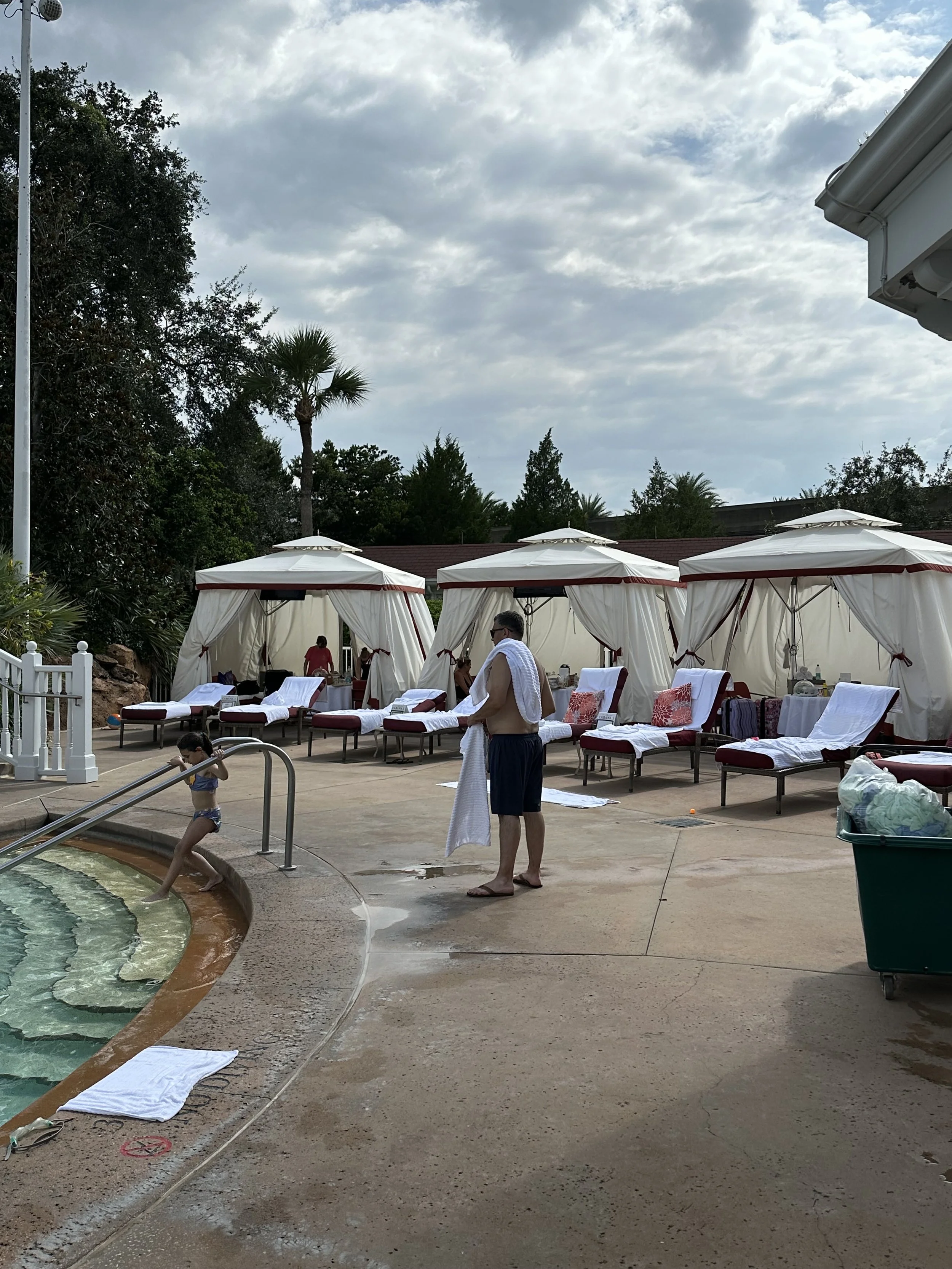 Poolside area with lounge chairs, some covered with towels or pillows, and private cabanas with curtains. A person in swim trunks and a towel is drying off, while a little girl in a swimsuit plays in the pool. The scene is outdoors with trees and a c
