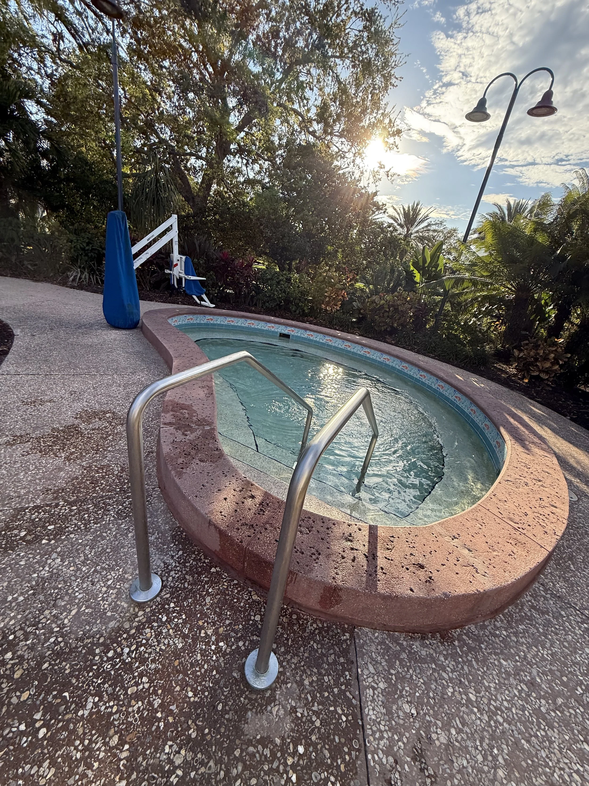 An outdoor hot tub with metal handrails sitting on a concrete patio surrounded by lush green trees and plants, with the sun shining through the trees and a clear sky with some clouds.