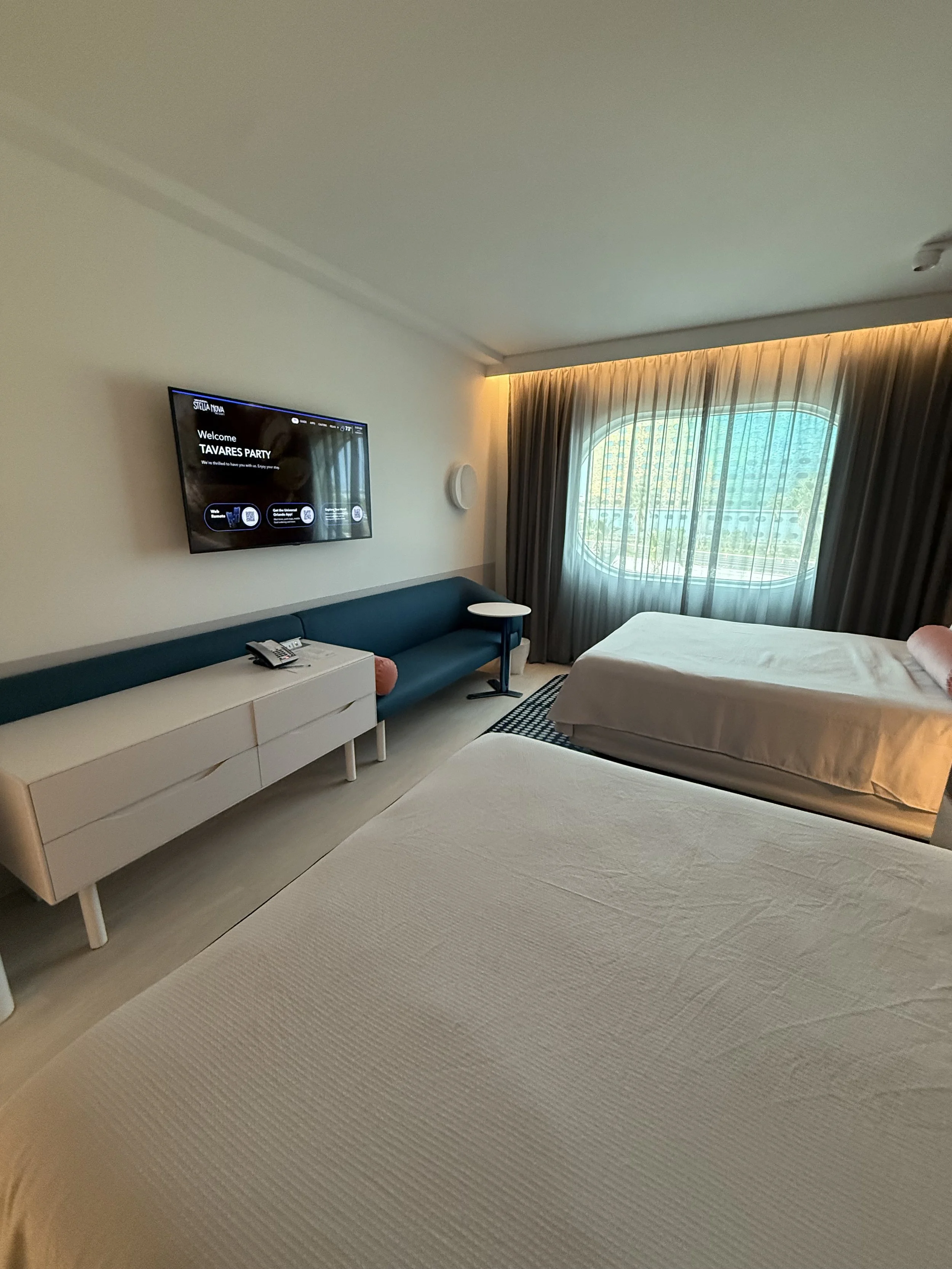 A hotel room with two beds, a teal sofa, a white dresser with a telephone, a TV on the wall, a side table, and a large window with sheer curtains.