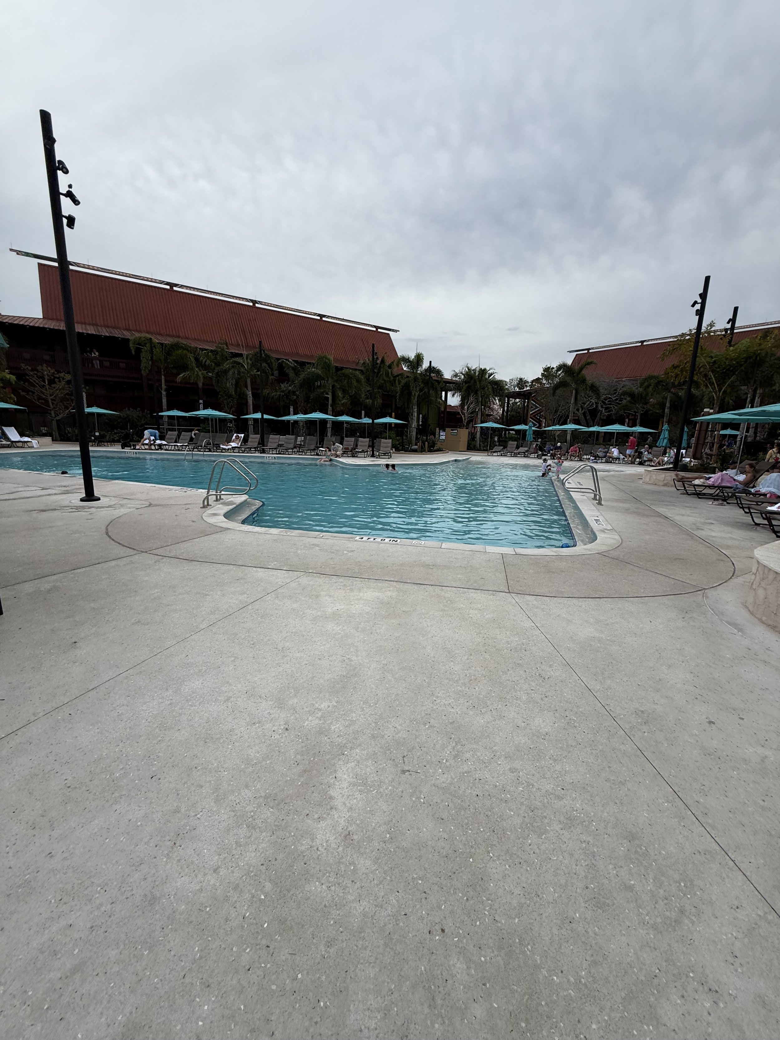A public swimming pool area with lounge chairs, blue umbrellas, and palm trees, under a cloudy sky.