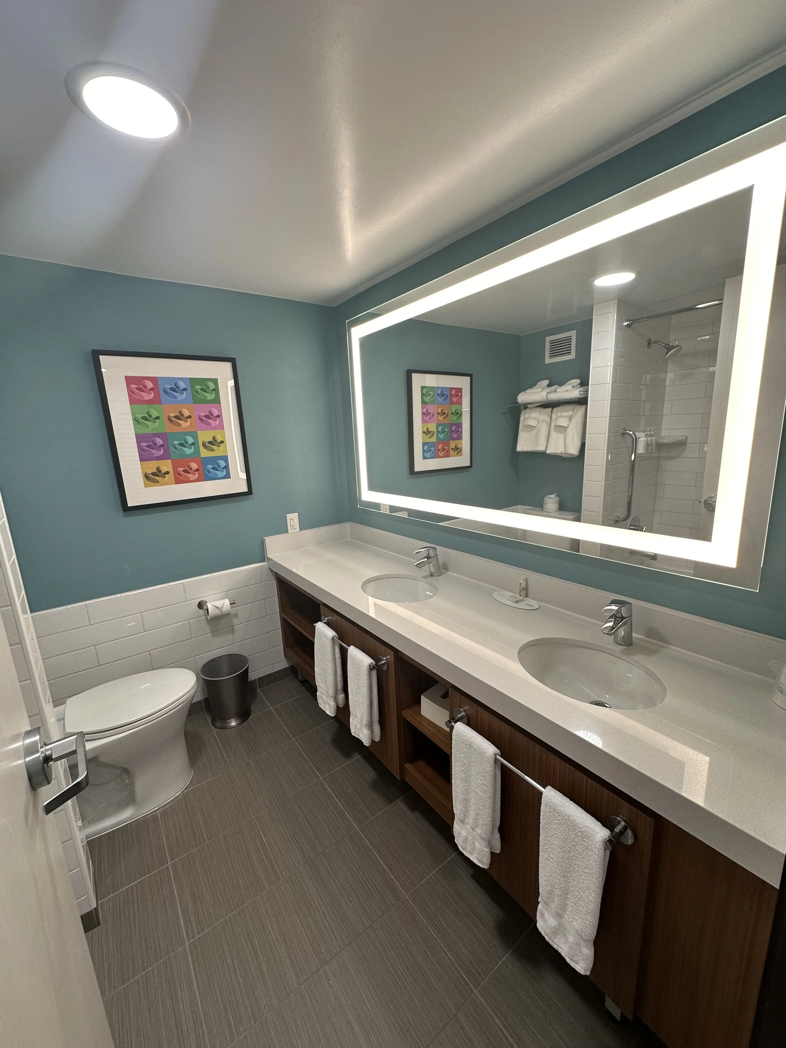Modern bathroom with double sinks, large illuminated mirror, toilet, framed colorful pop art on teal walls, white tiled shower area, and brown flooring.