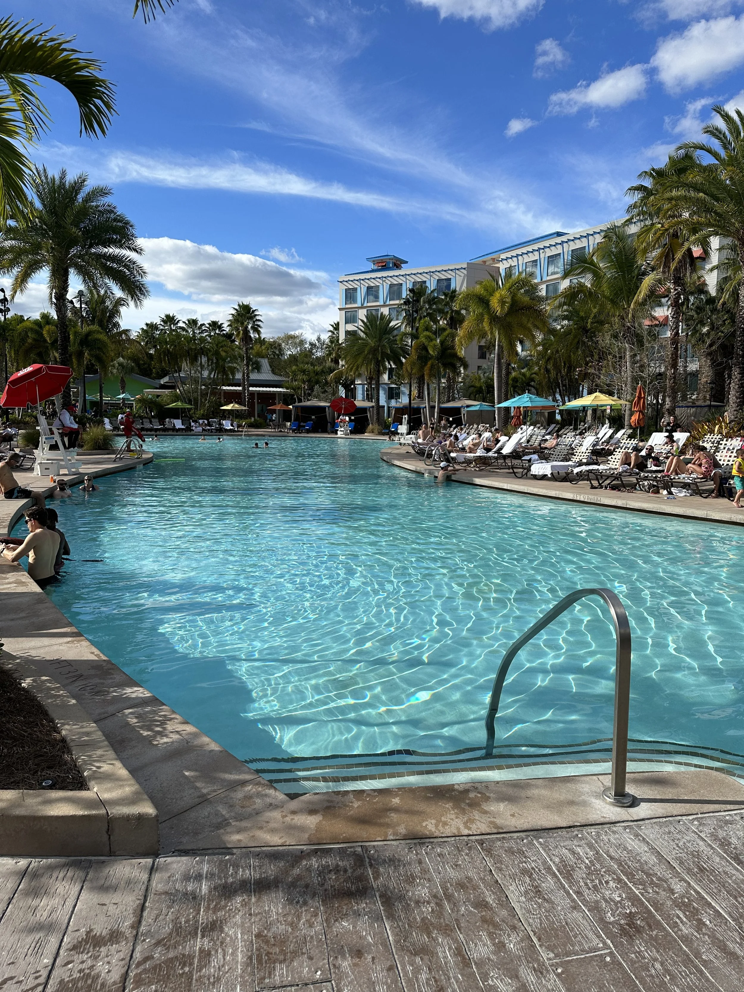 A hotel swimming pool area with people swimming and sunbathing, surrounded by palm trees and umbrellas under a partly cloudy blue sky.
